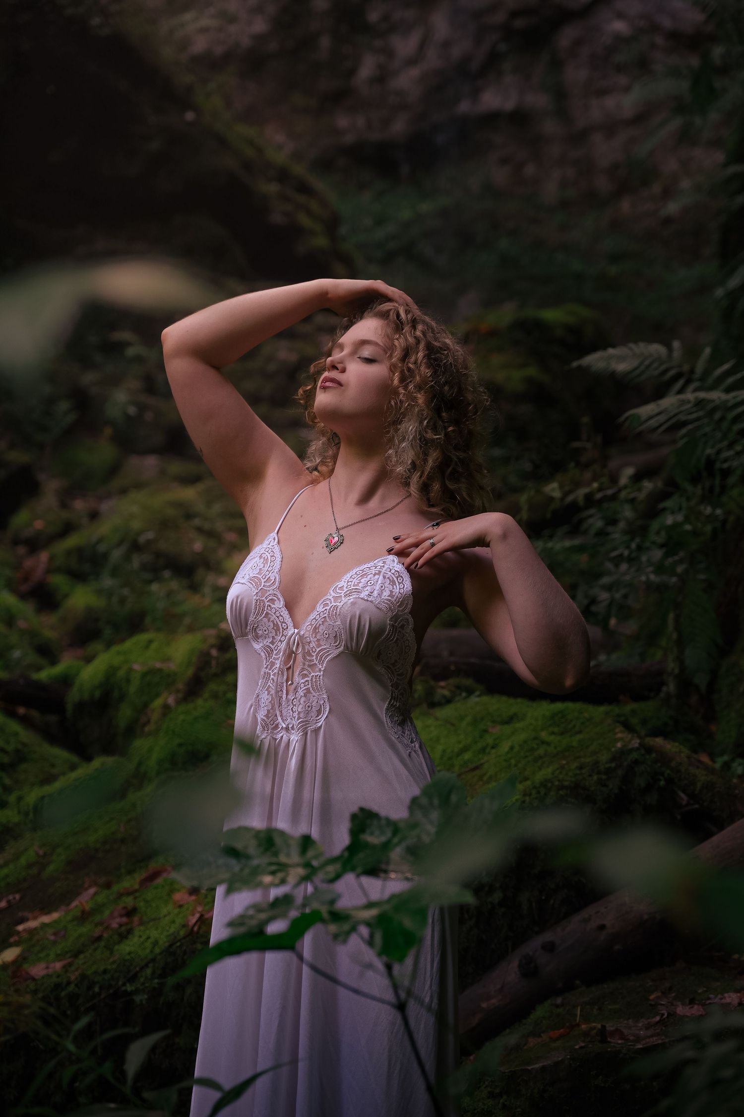 Woman in white lace dress poses gracefully among moss-covered rocks in a lush forest setting.
