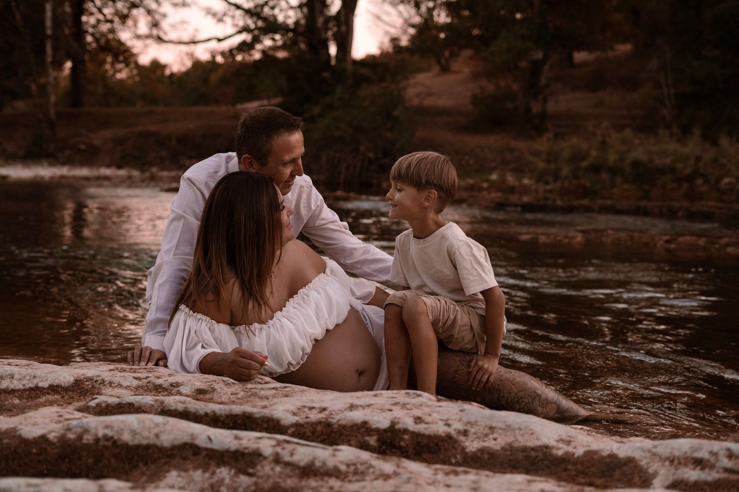 Pregnant woman sits with partner and young boy by a serene river at sunset during a family maternity photo session.