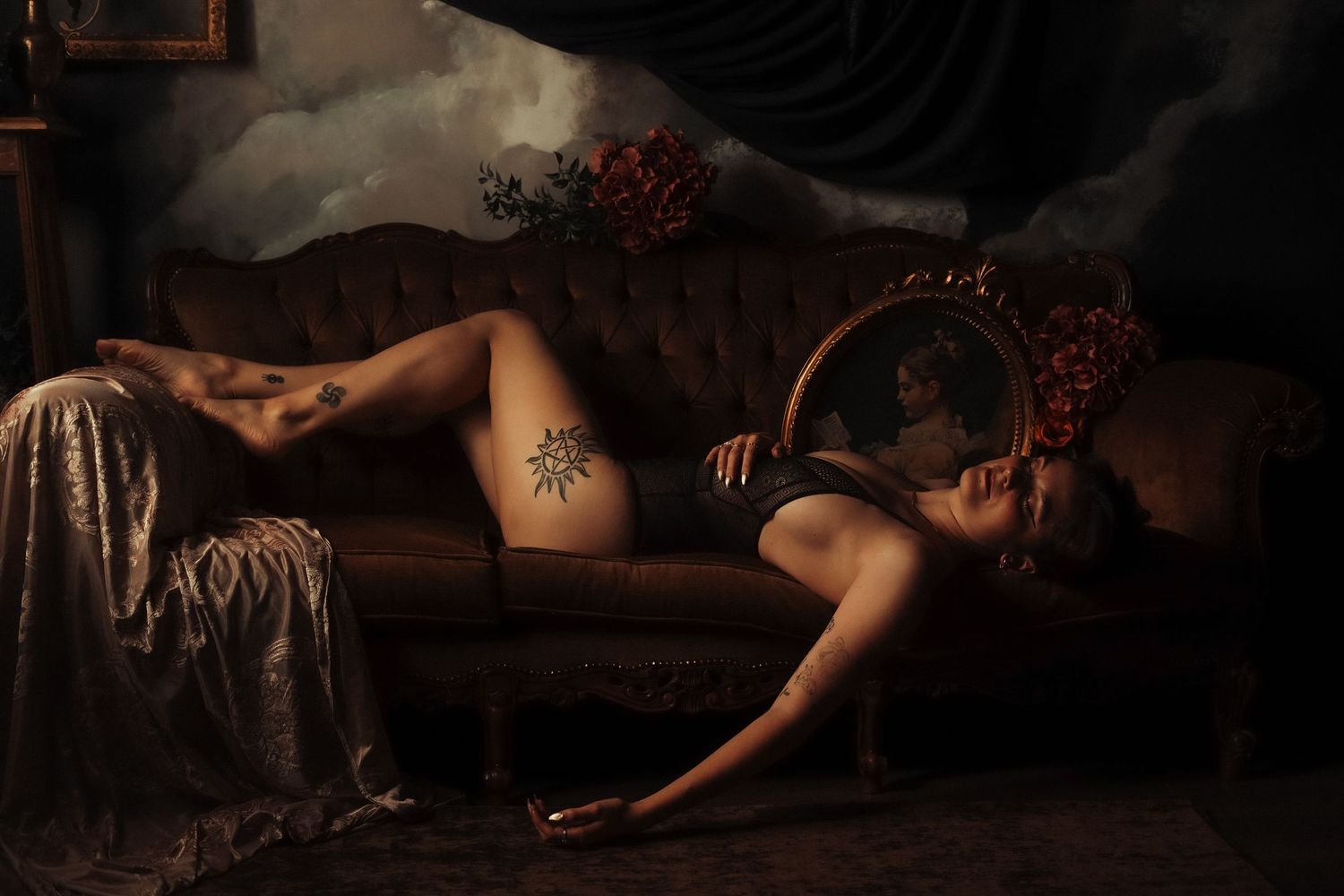 Tattooed woman reclining on vintage sofa in artistic dark boudoir scene with floral decor and mirror.
