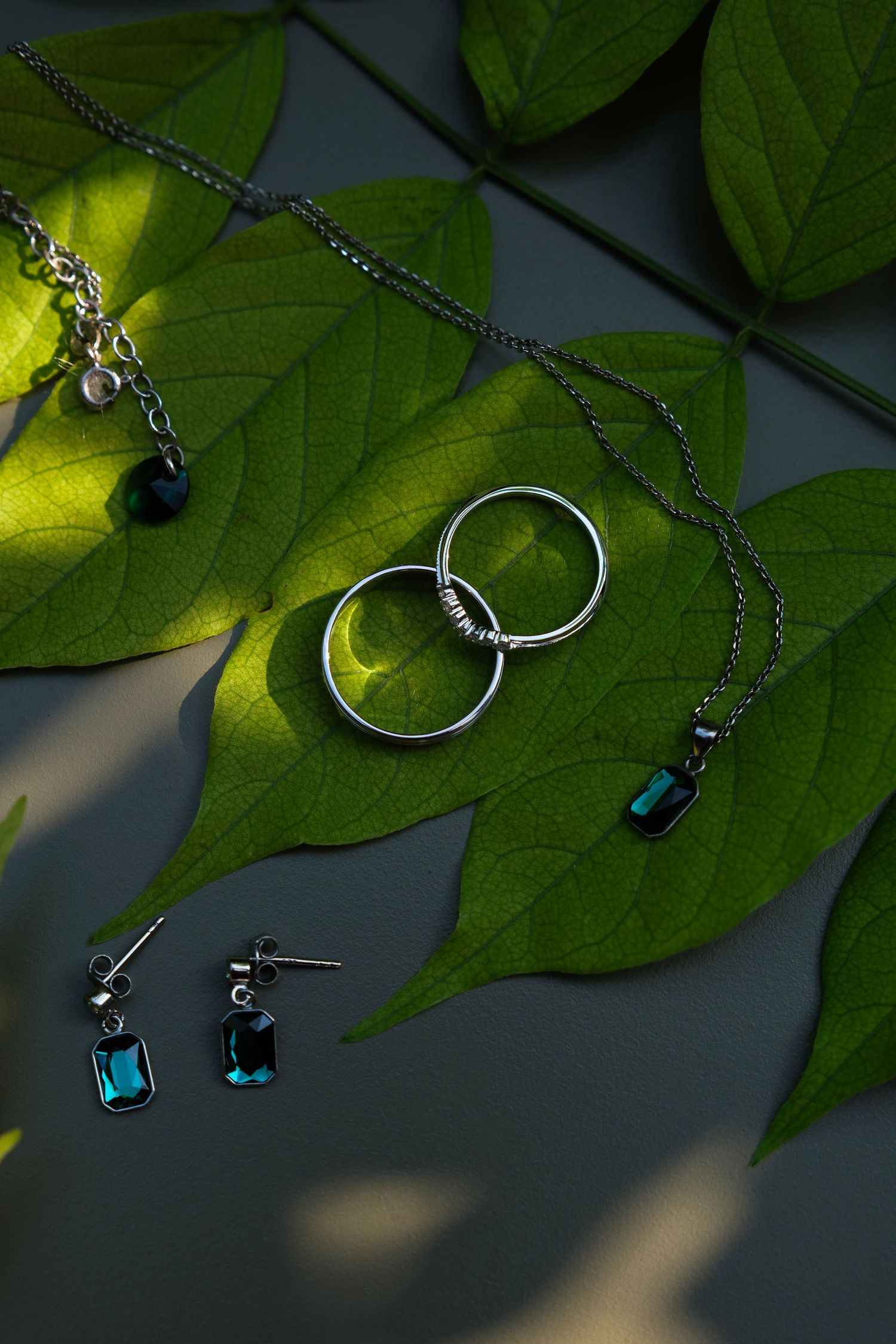 Wedding rings, earrings, and necklaces with teal gemstones arranged on green leaves.