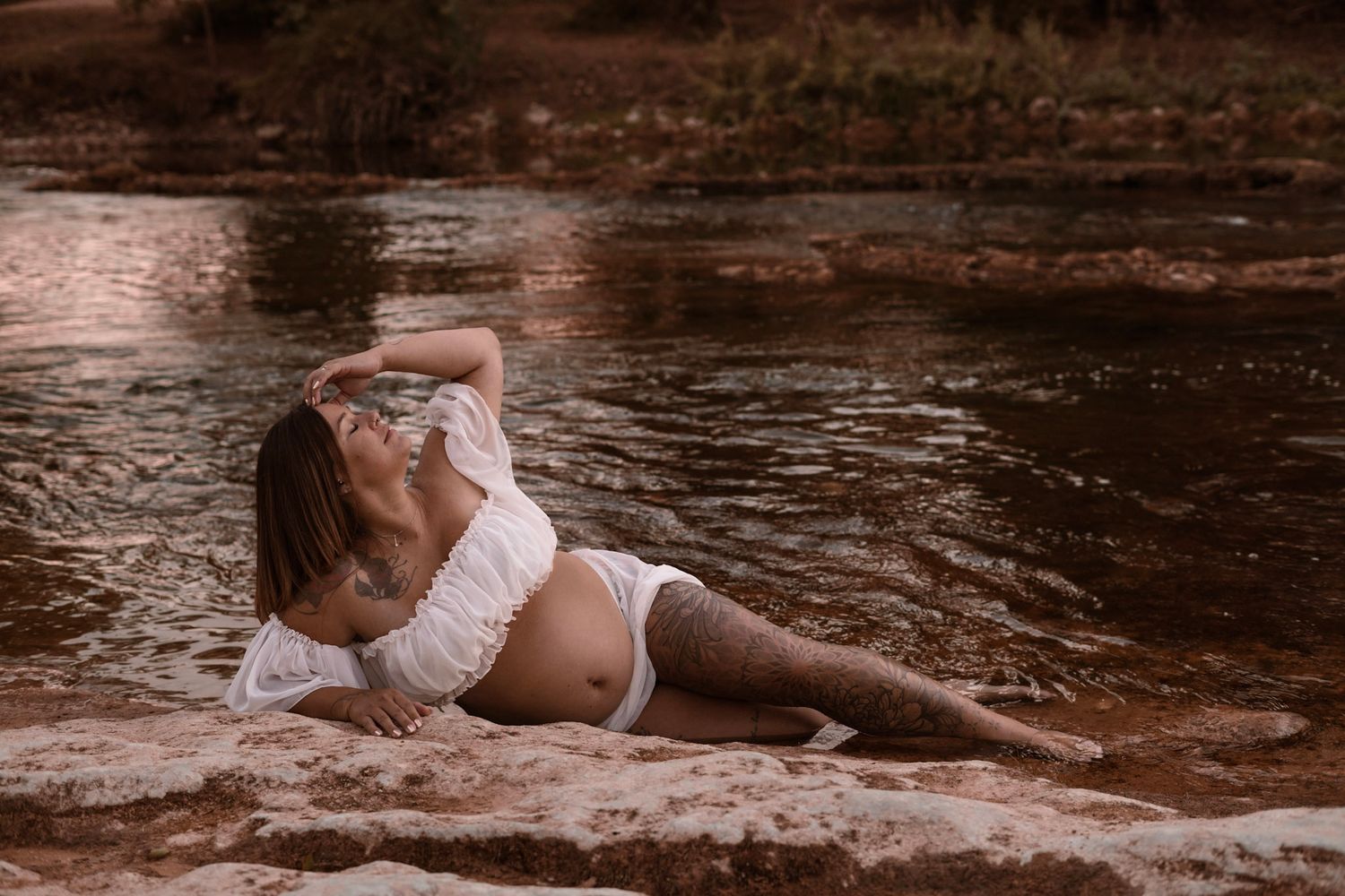 Pregnant woman in white lace dress posing gracefully on rocks by a serene river in warm tones.
