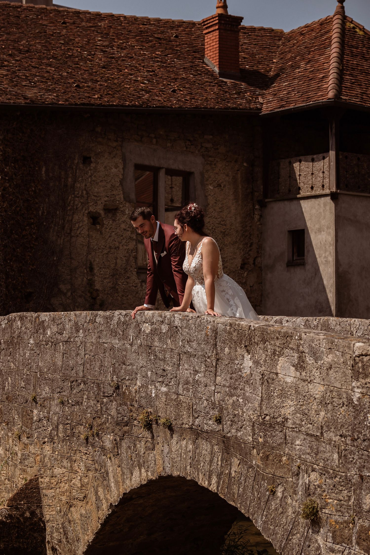 Bride and groom walk hand in hand across a rustic stone bridge near a historic building.
