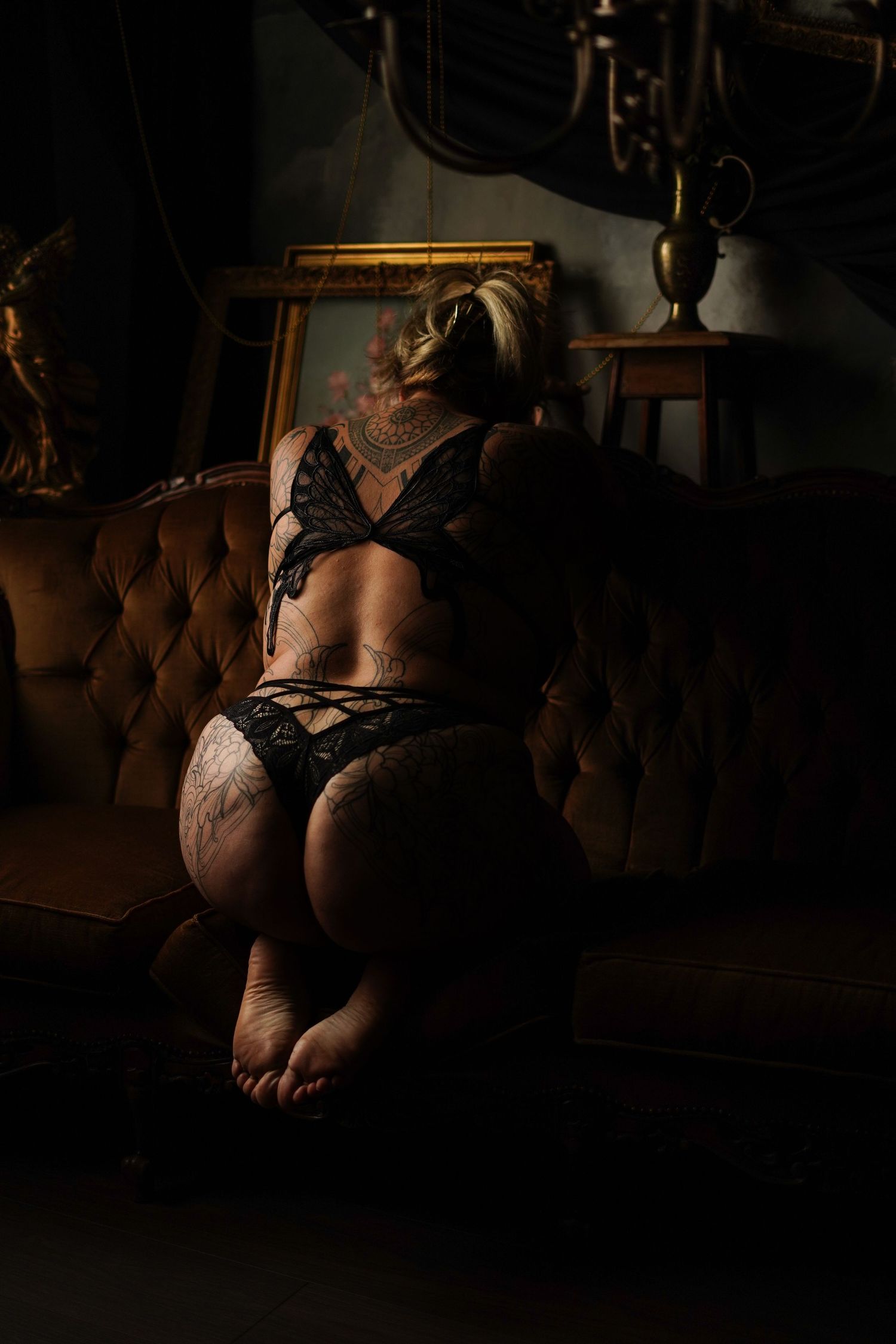 A tattooed woman in black lace lingerie poses on a vintage velvet sofa in moody boudoir setting.