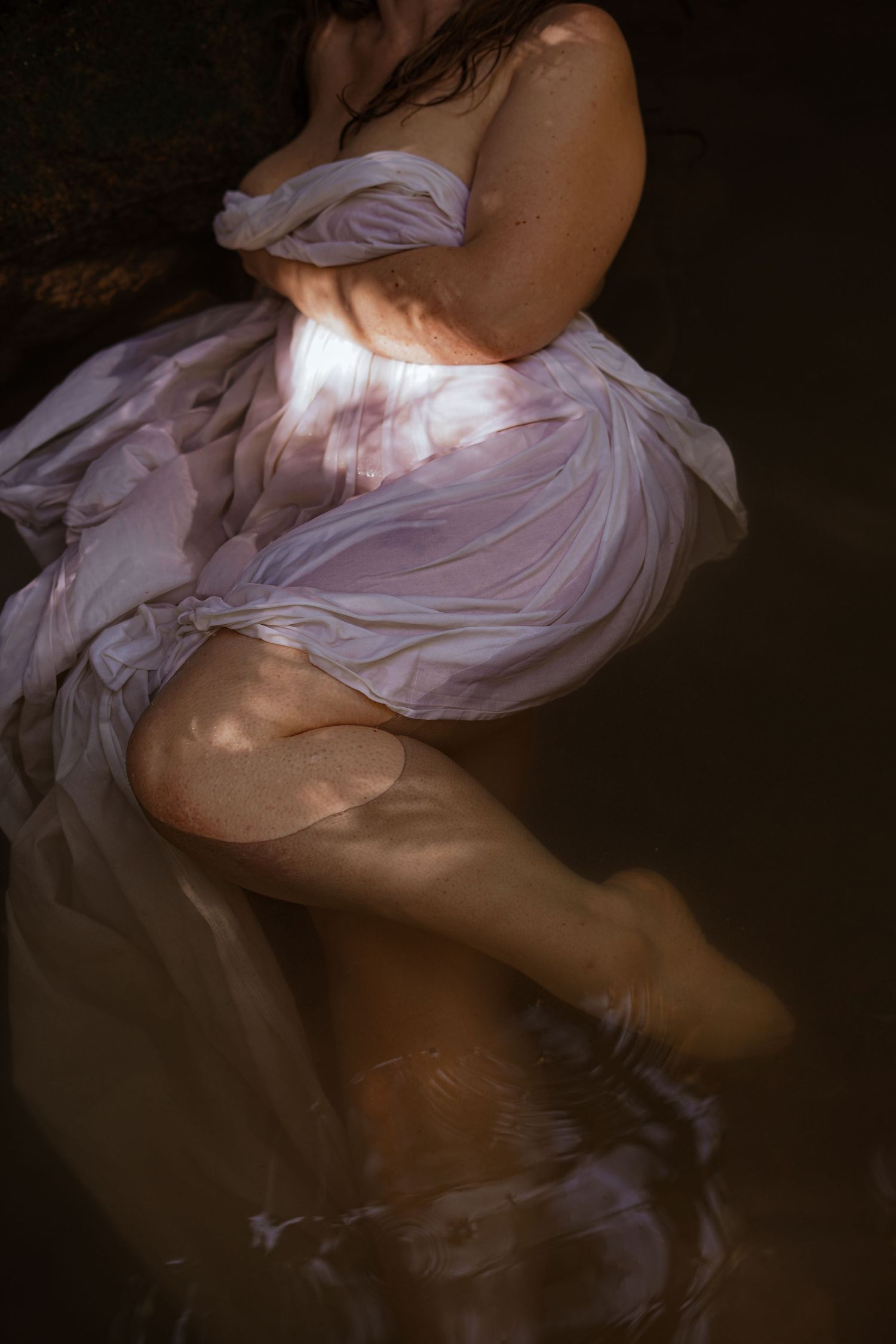 Woman in flowing white sheer dress standing in water with soft natural lighting.