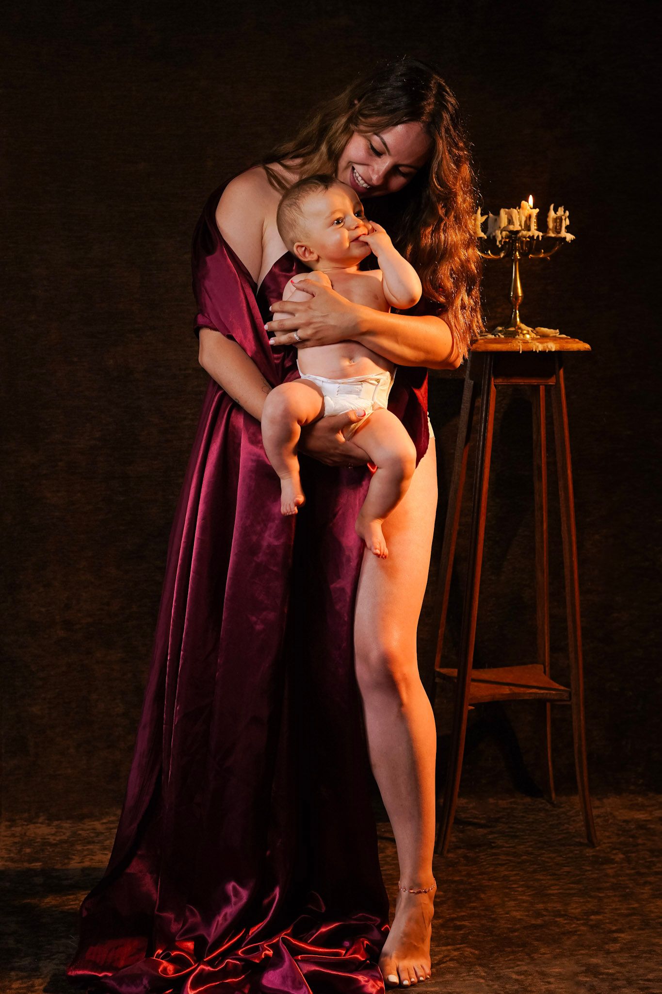 Woman in burgundy gown holding smiling baby near candlelit table in dramatic studio portrait.