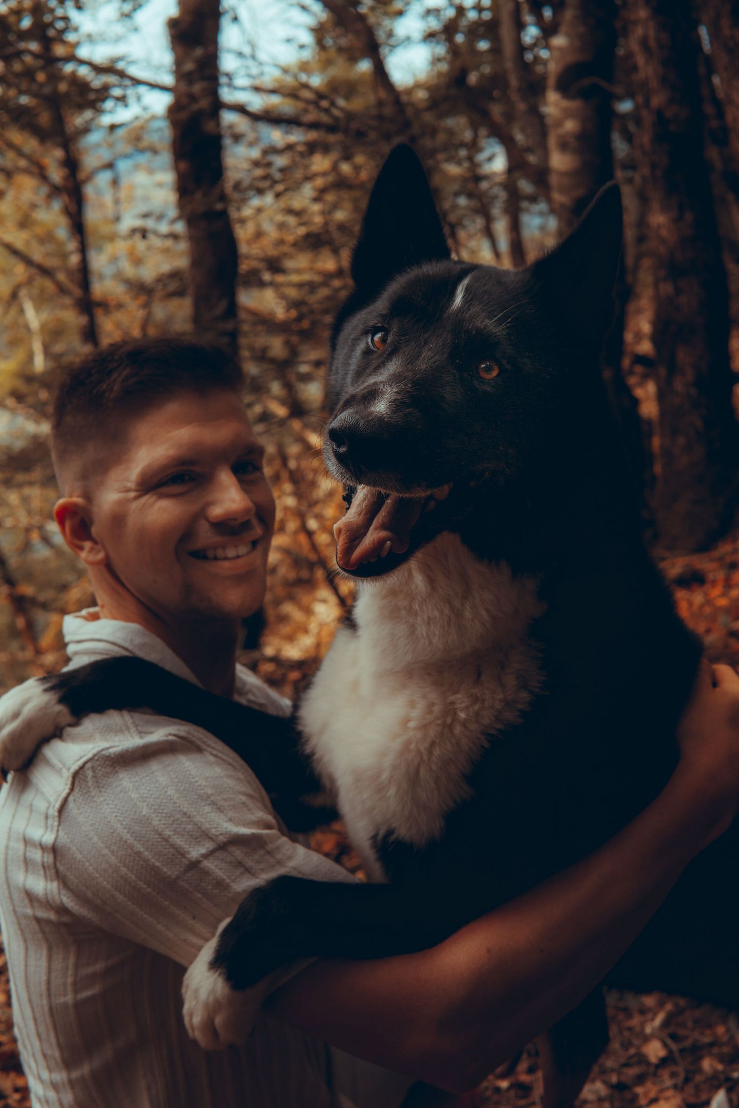Smiling man holding happy black and white dog in colorful autumn forest setting.