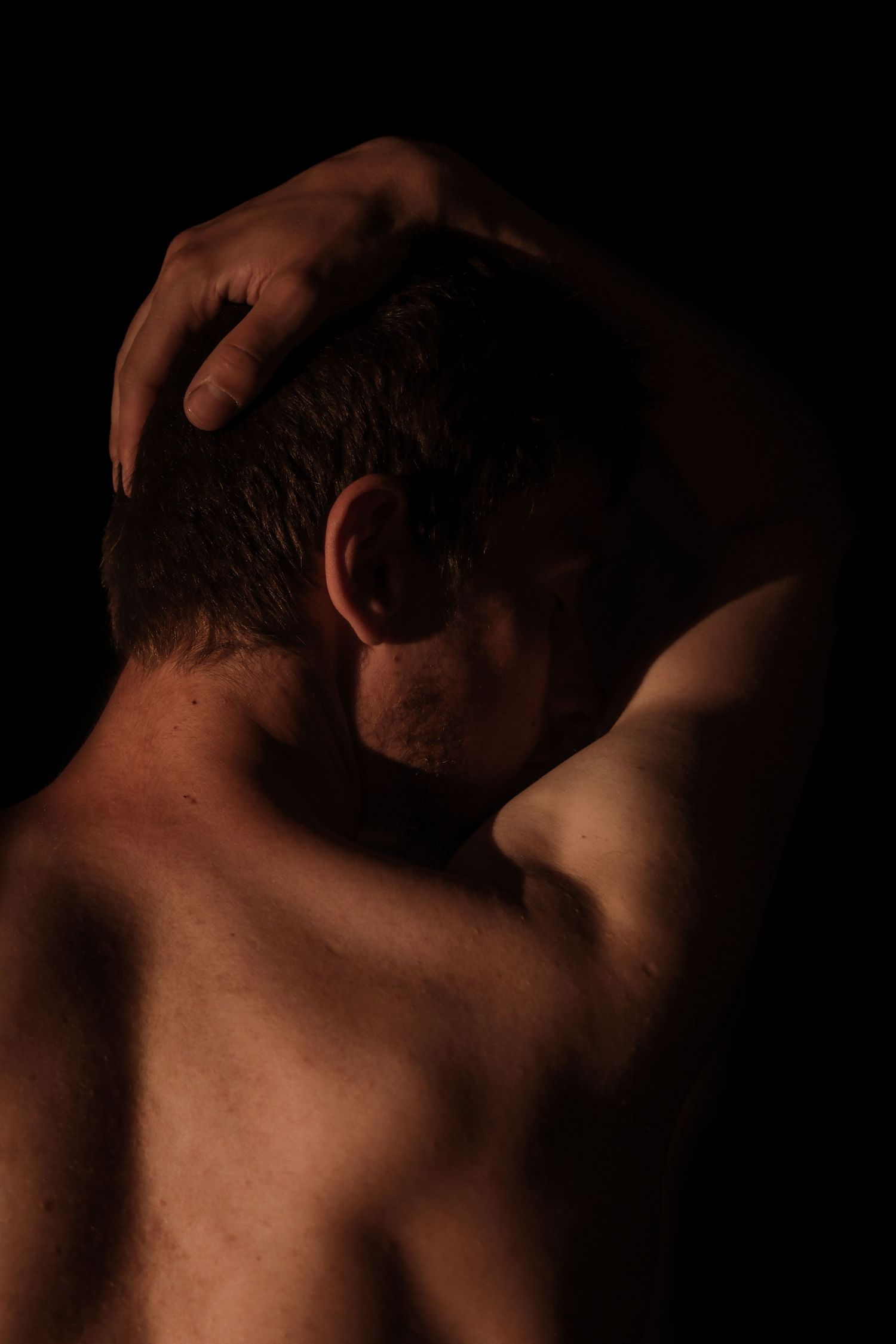 Shirtless man photographed from behind with hand on head against dark background.