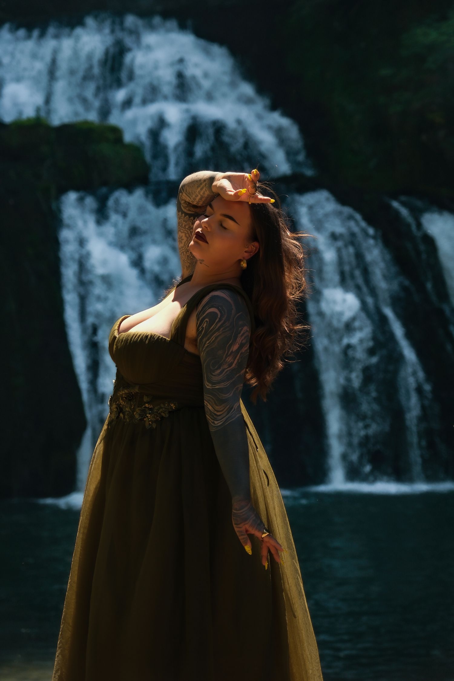 Tattooed woman in olive gown poses before majestic waterfall backdrop.