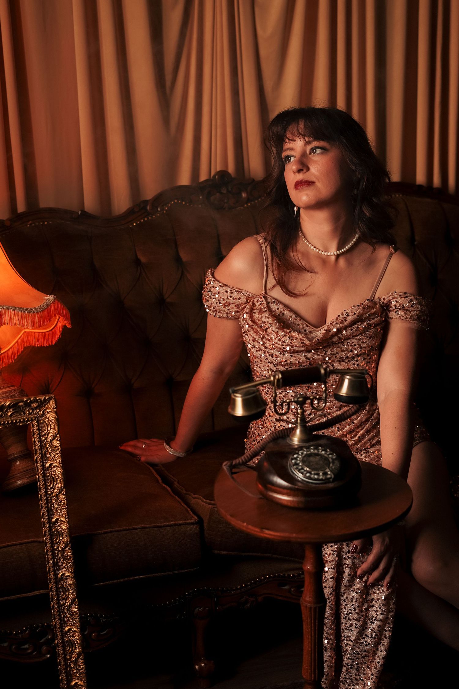 Woman in sequin dress sitting on vintage sofa next to retro rotary telephone.