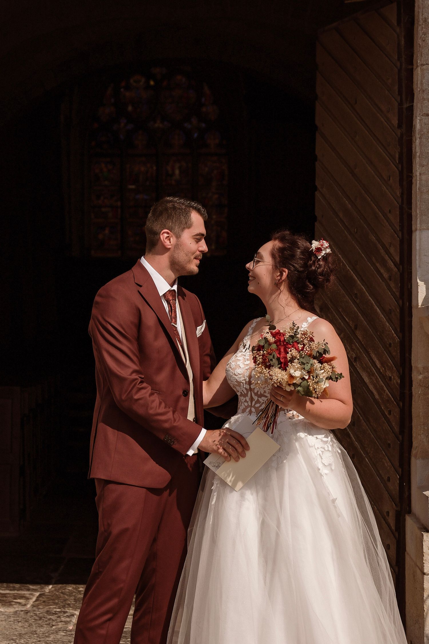Bride holding bouquet and groom in burgundy suit smiling at each other outside church.