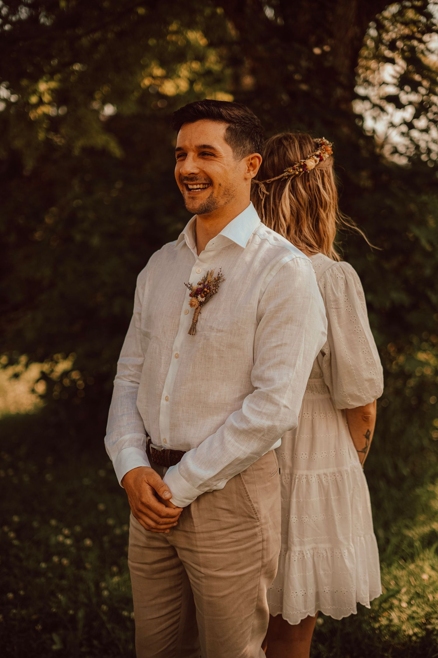 Smiling groom in white linen shirt with boutonniere stands outdoors in golden hour light.