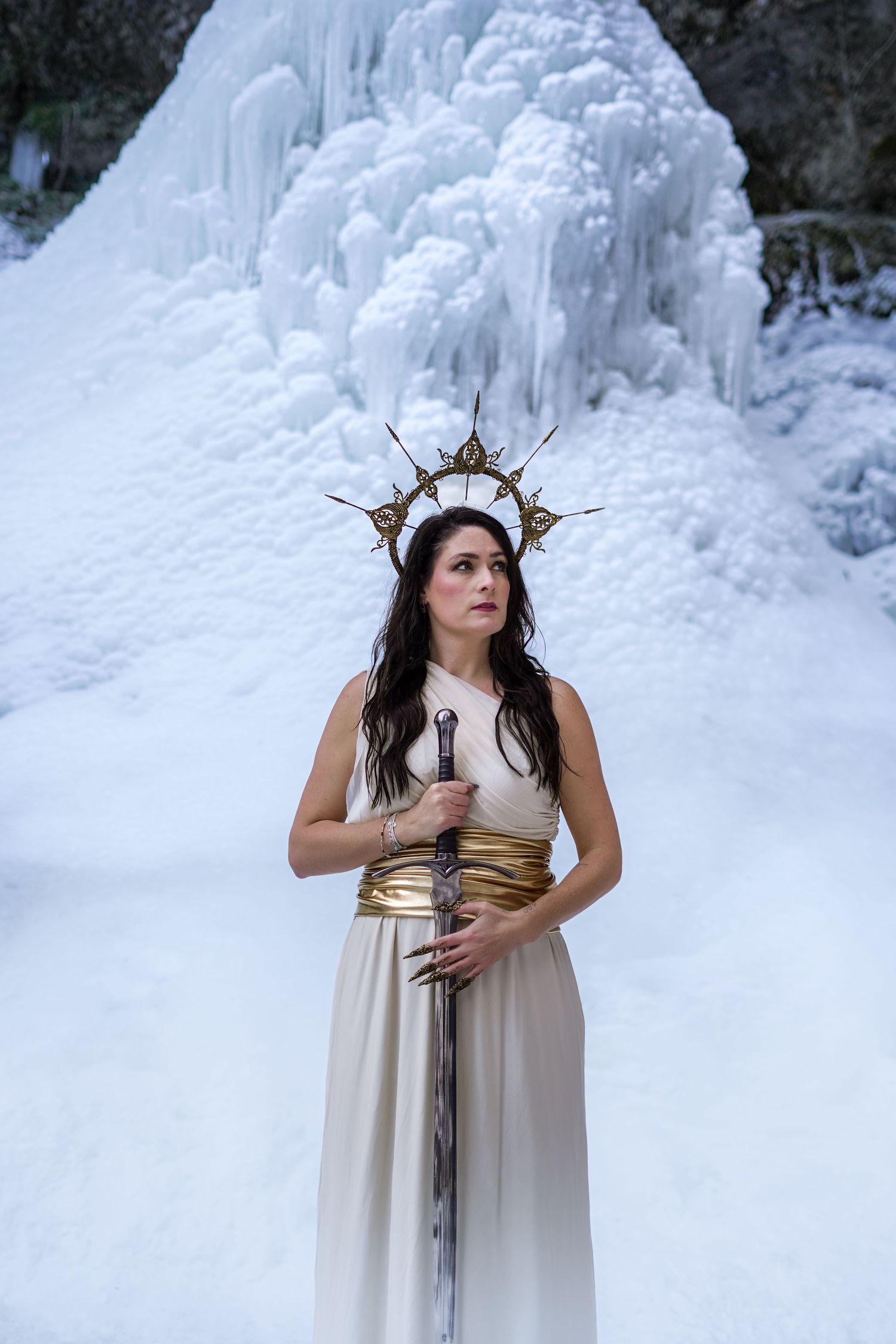 Woman in white gown with gold crown holding sword before icy waterfall.