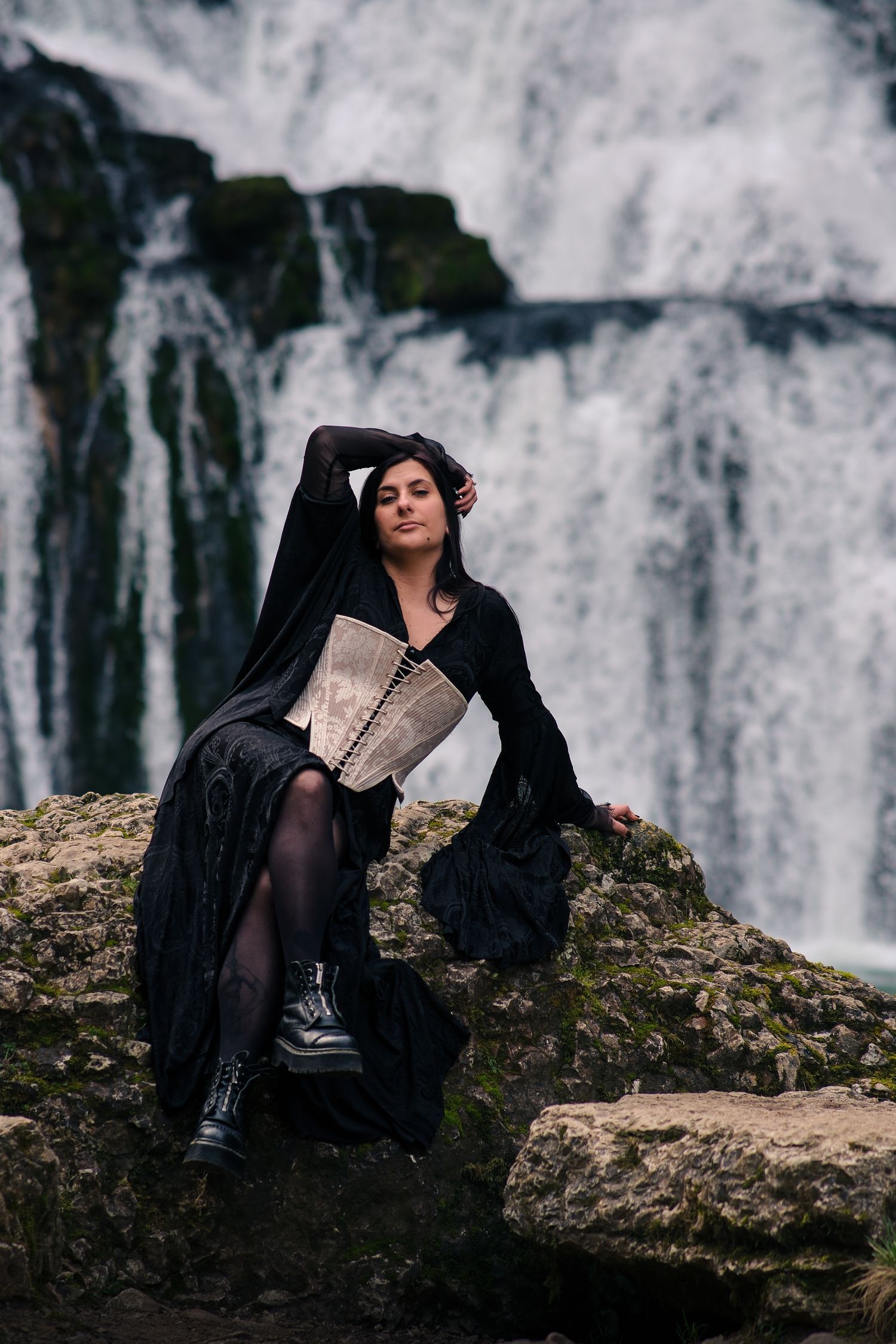 Woman in black gothic dress with corset posing on rocks before a dramatic waterfall.
