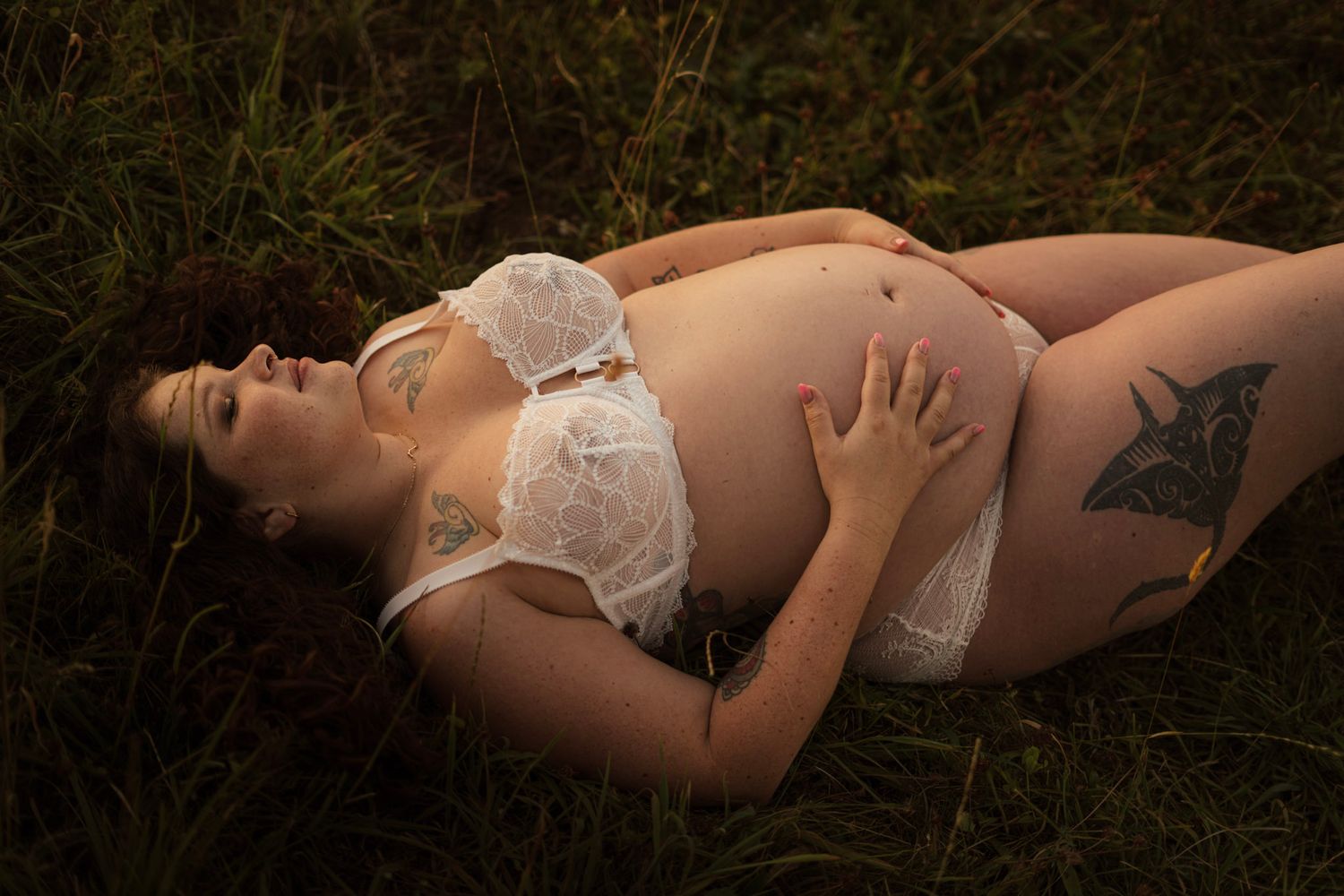 Pregnant woman in white lace lingerie lying in grass, showcasing baby bump with tattoo visible on hip.