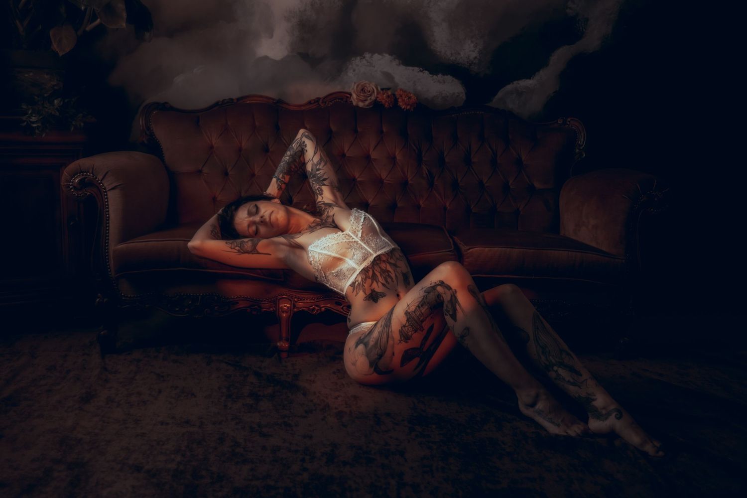 Tattooed woman in white lingerie lying on brown velvet sofa in dramatic dark setting.