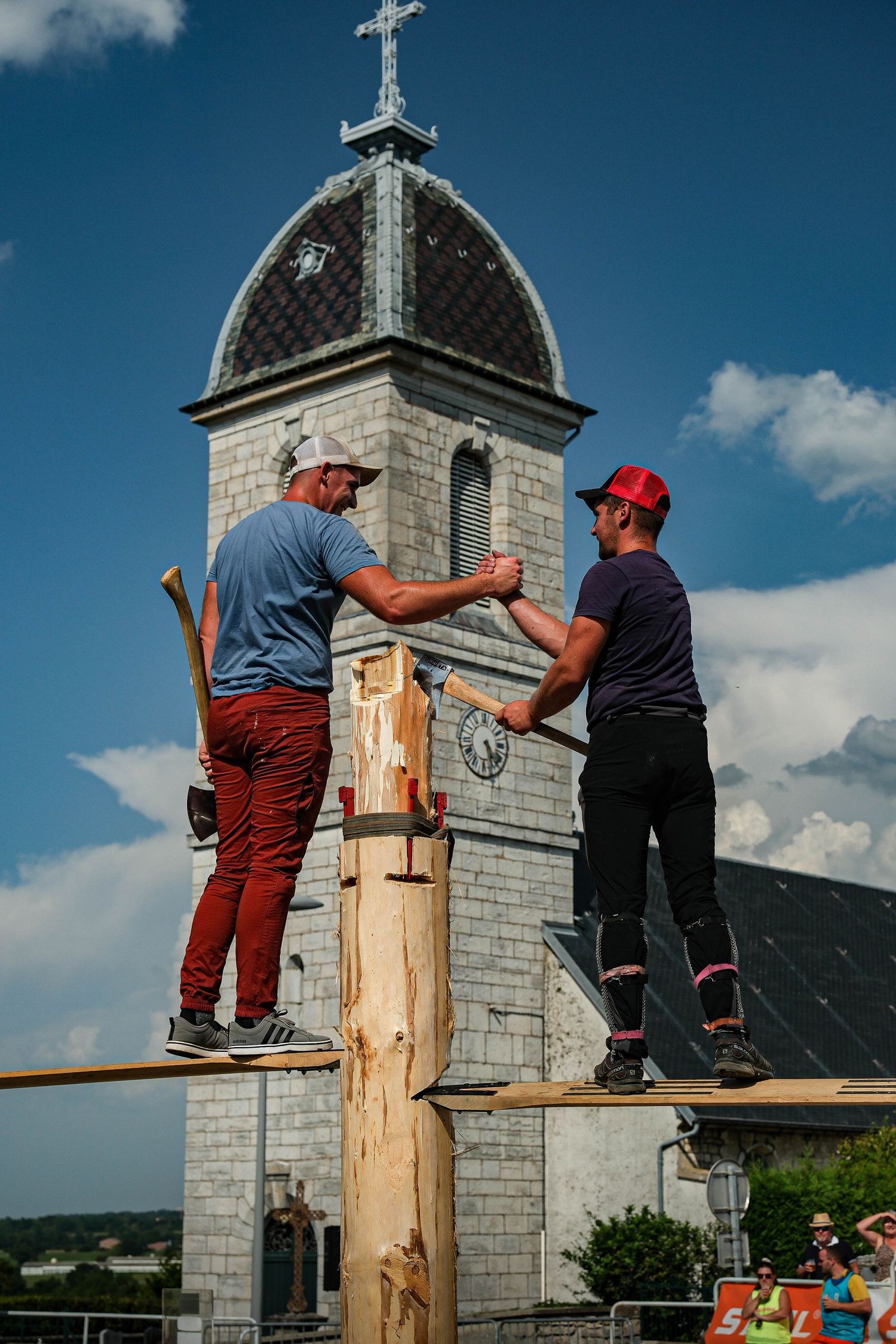 Two men working atop wooden pole with historic church steeple in background during outdoor event.
