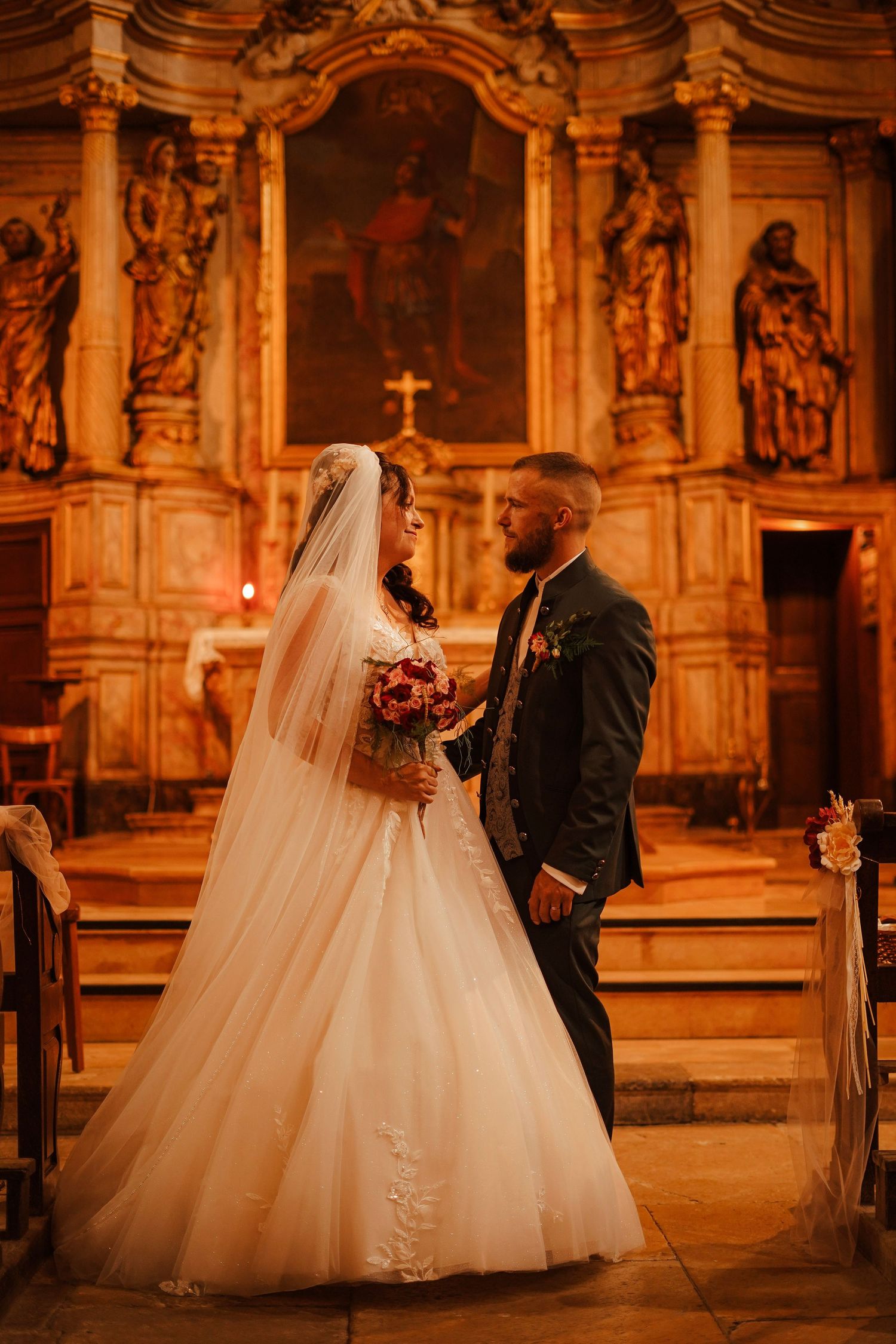 Bride and groom facing each other in front of golden baroque church altar holding red bouquet.