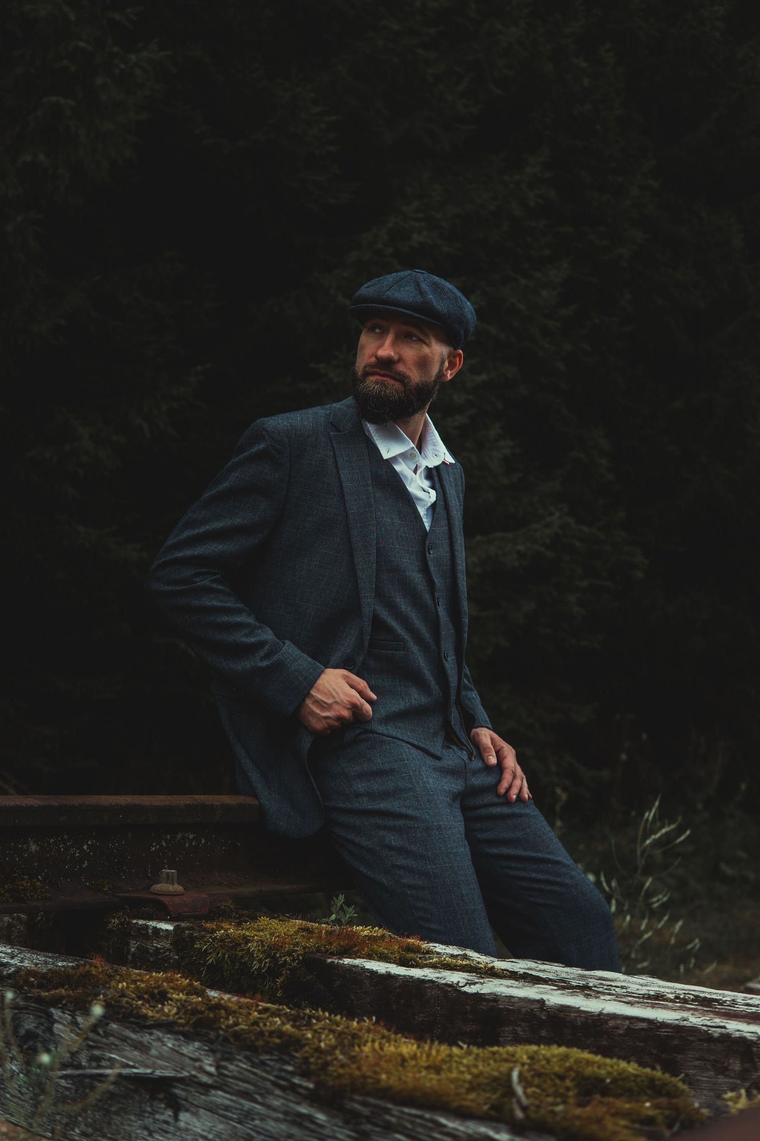 A bearded man in a vintage grey three-piece suit and flat cap sits on logs in a dark forest setting.