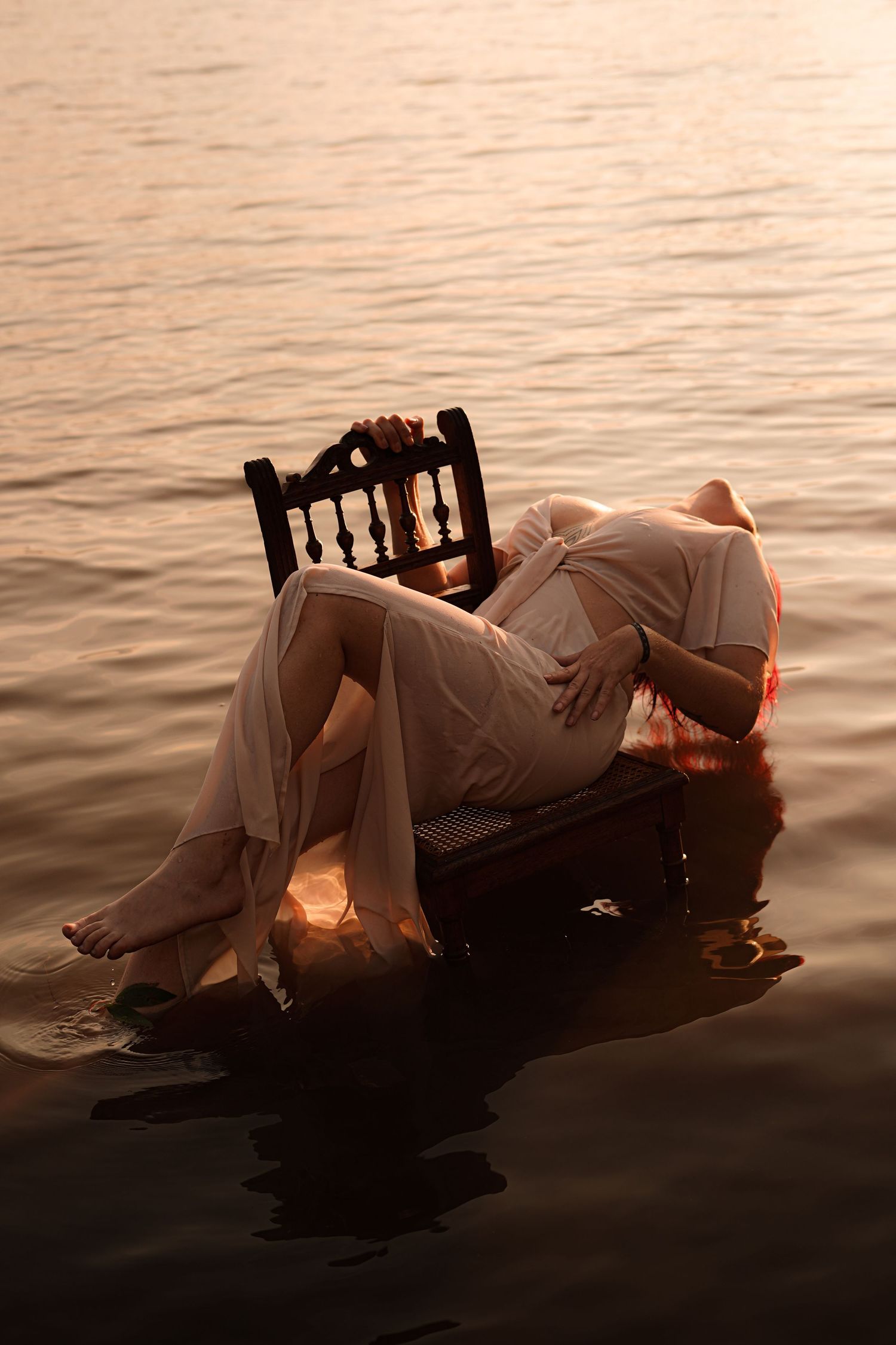 A woman in a white dress drapes over a wooden chair floating on calm golden sunset-lit water.