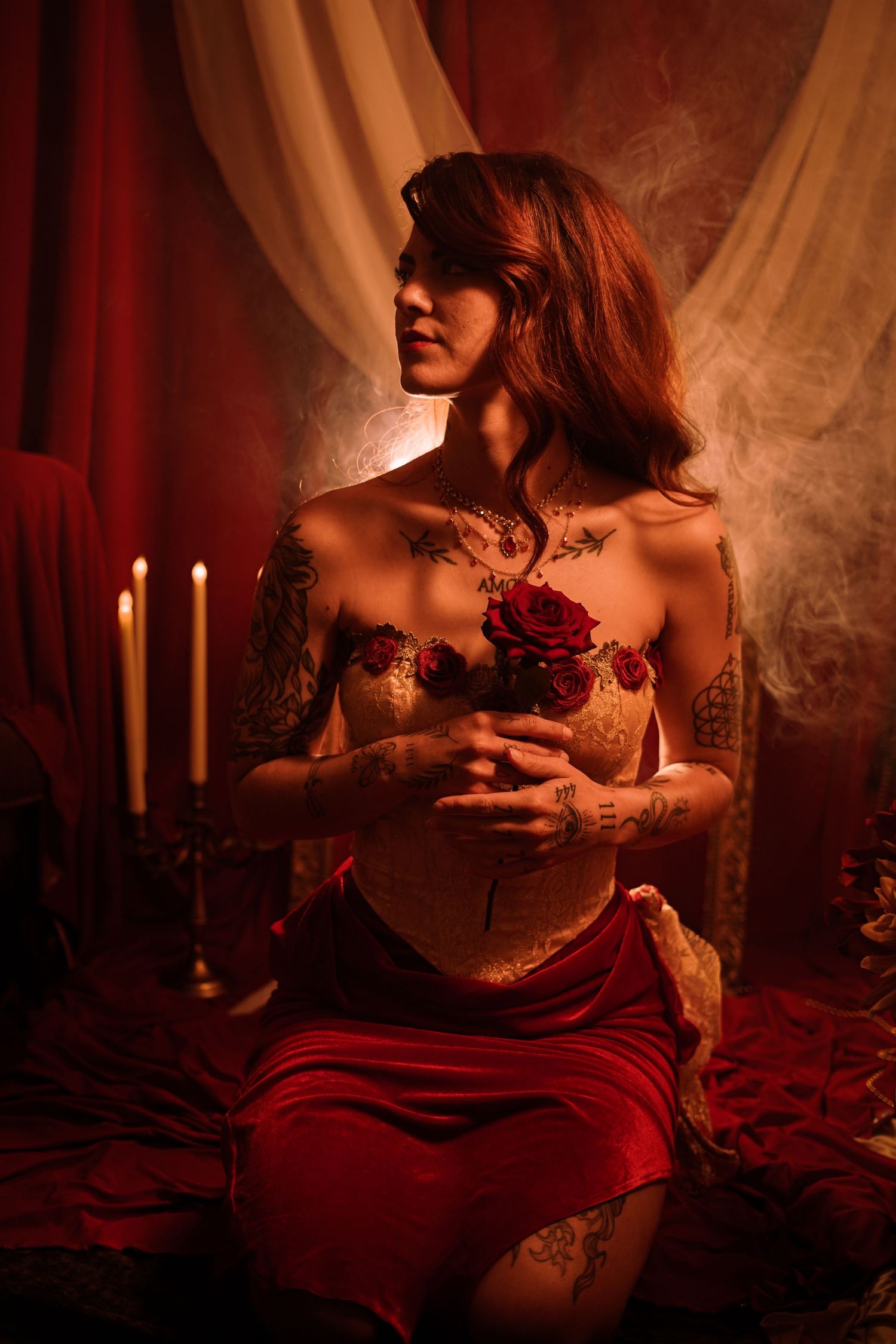 Tattooed woman in lace lingerie holding red roses beside candles in dramatic red-lit setting.