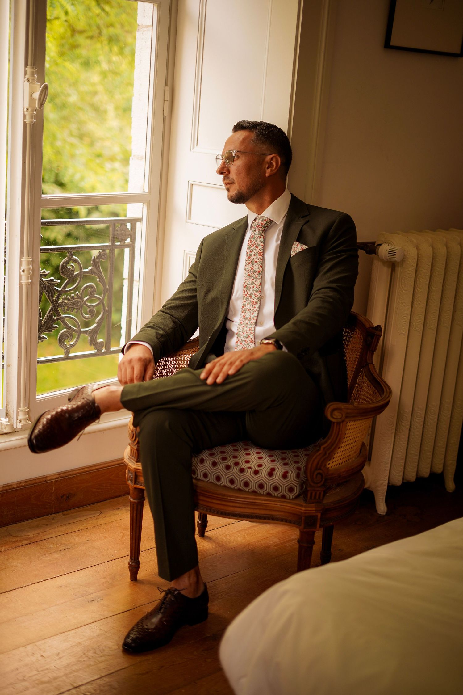 Elegant groom in dark suit sitting in vintage armchair gazing out sunlit window.