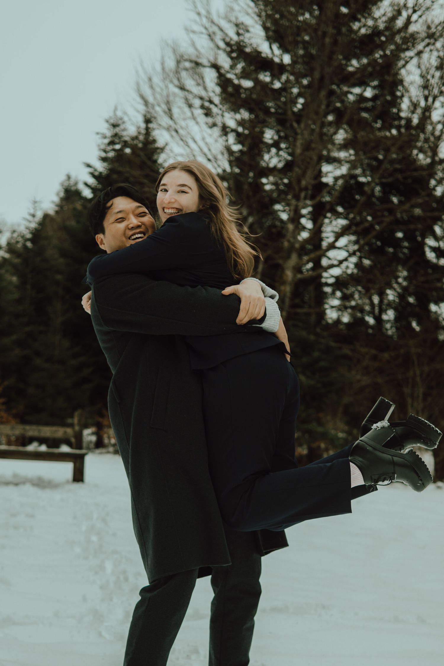 Couple embracing in snowy winter landscape, man lifting woman in black coats with pine trees behind.
