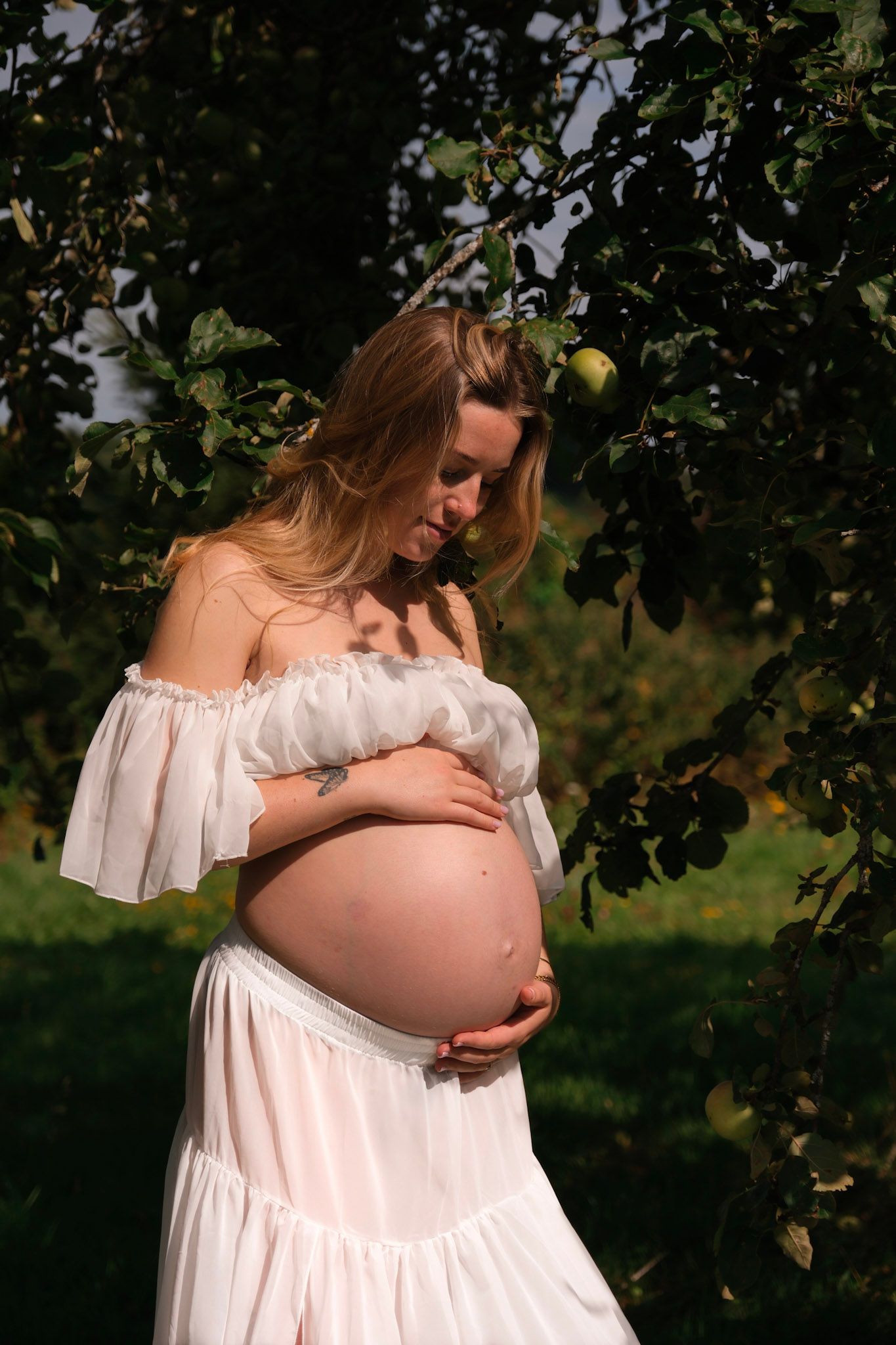 Pregnant woman in white off-shoulder dress cradling baby bump outdoors near apple tree in sunlight.