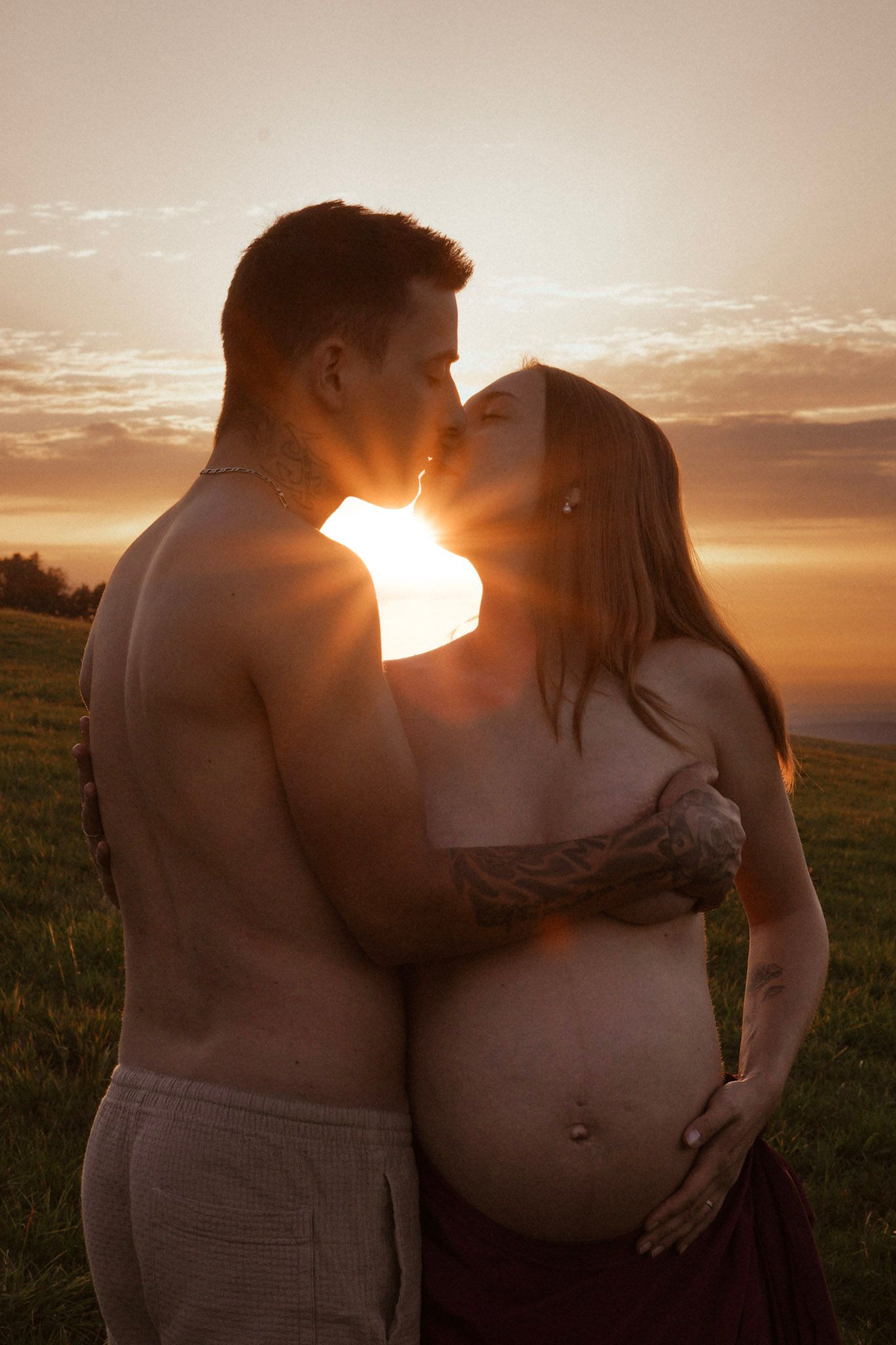 Pregnant couple sharing a kiss at golden sunset on a green hillside, man shirtless with tattoos, woman cradling baby bump.