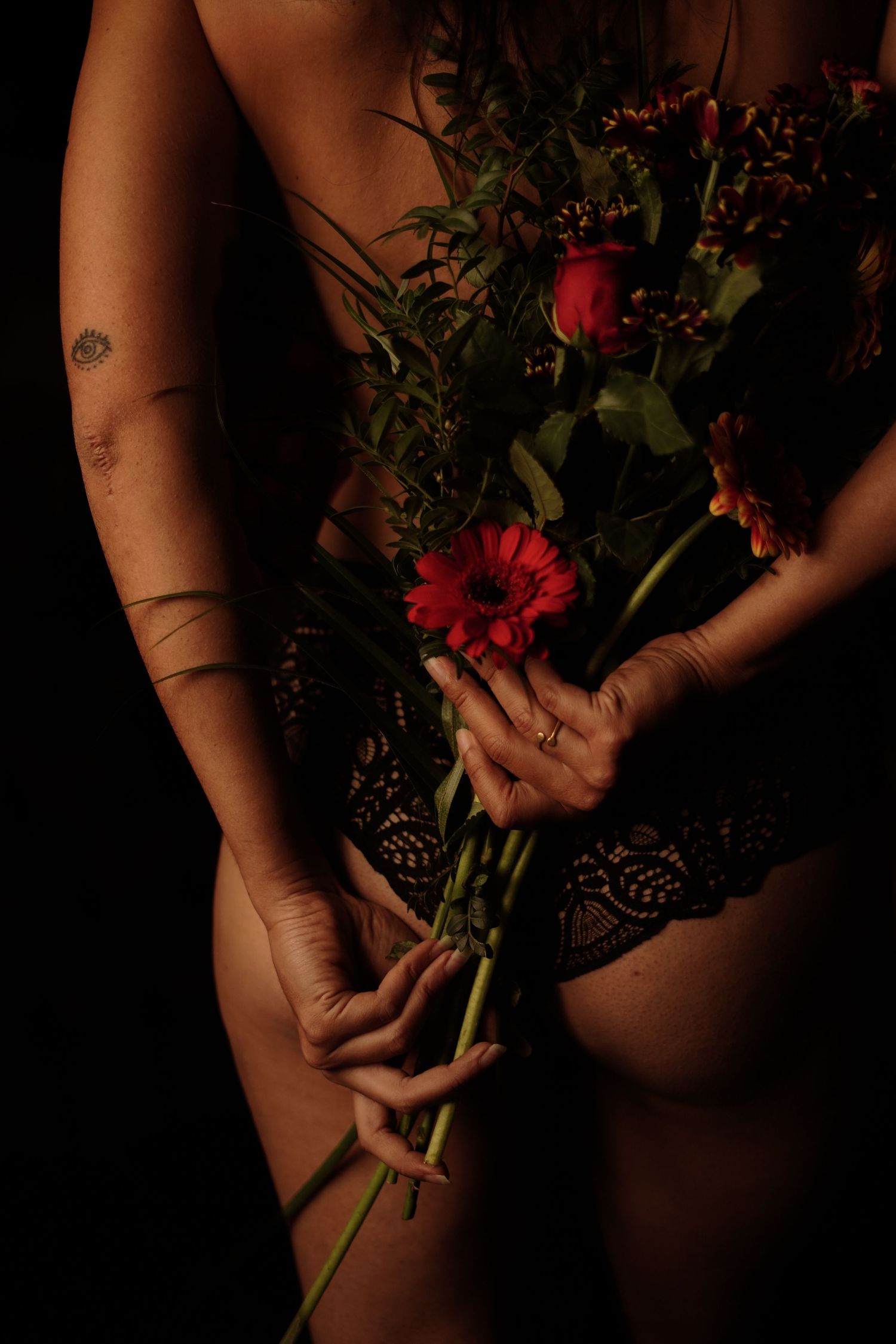 Woman in black lace lingerie holding bouquet of red flowers behind her back with tattoos visible.