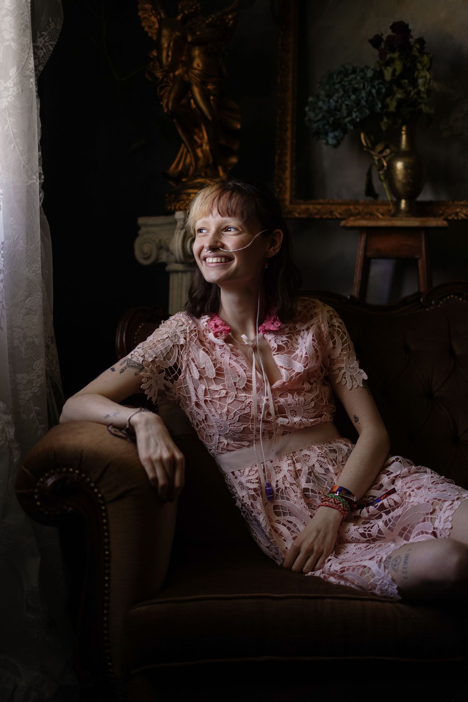 Smiling woman in floral pink lace dress relaxes in vintage armchair.