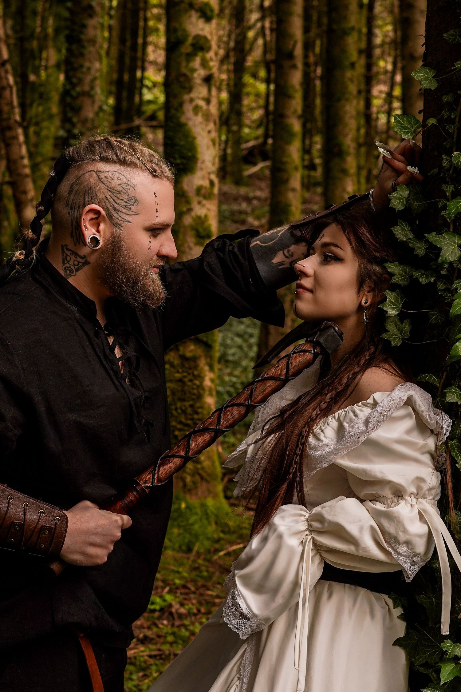 Viking-costumed couple in dramatic forest setting, man with facial tattoos and woman in white dress.