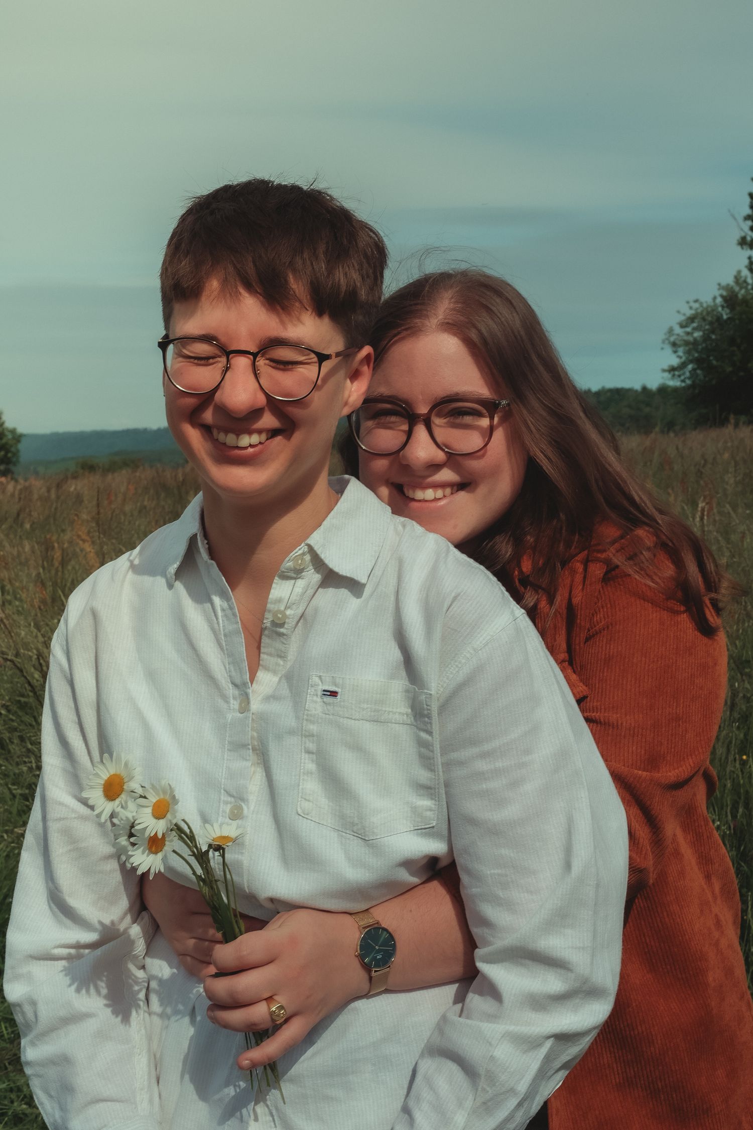 A smiling couple wearing glasses embraces in a meadow, one holding a bouquet of white daisies.