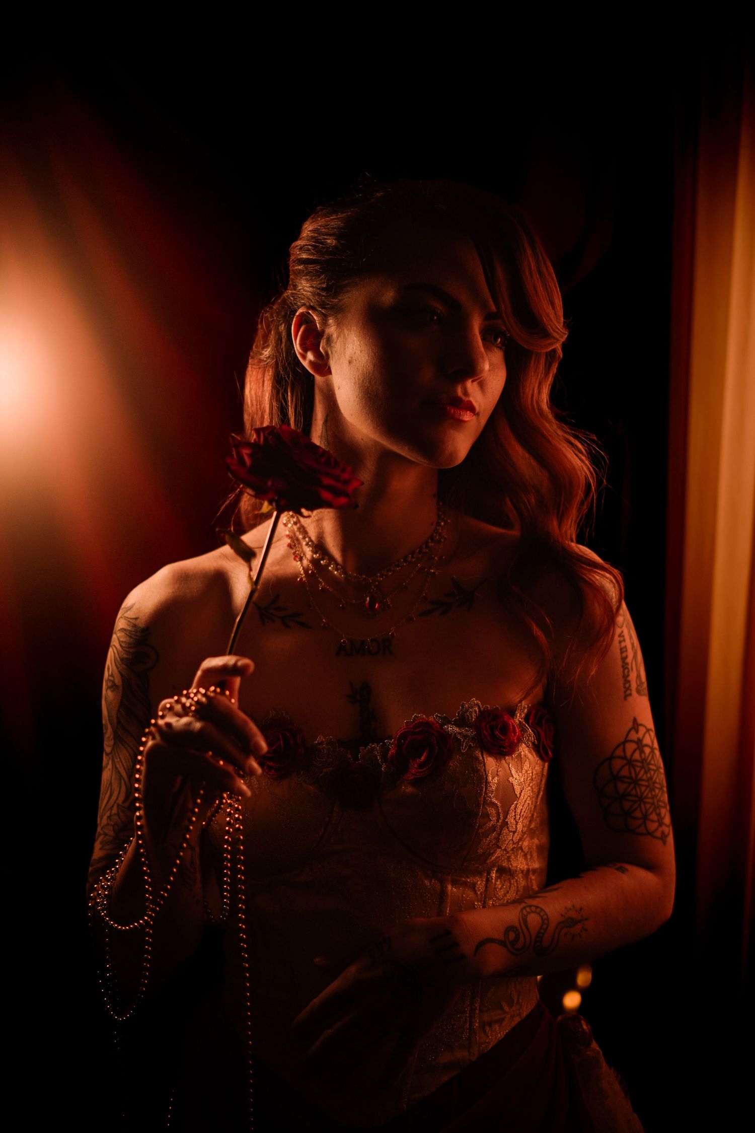 Tattooed woman holds red rose and pearls in warm candlelit portrait.
