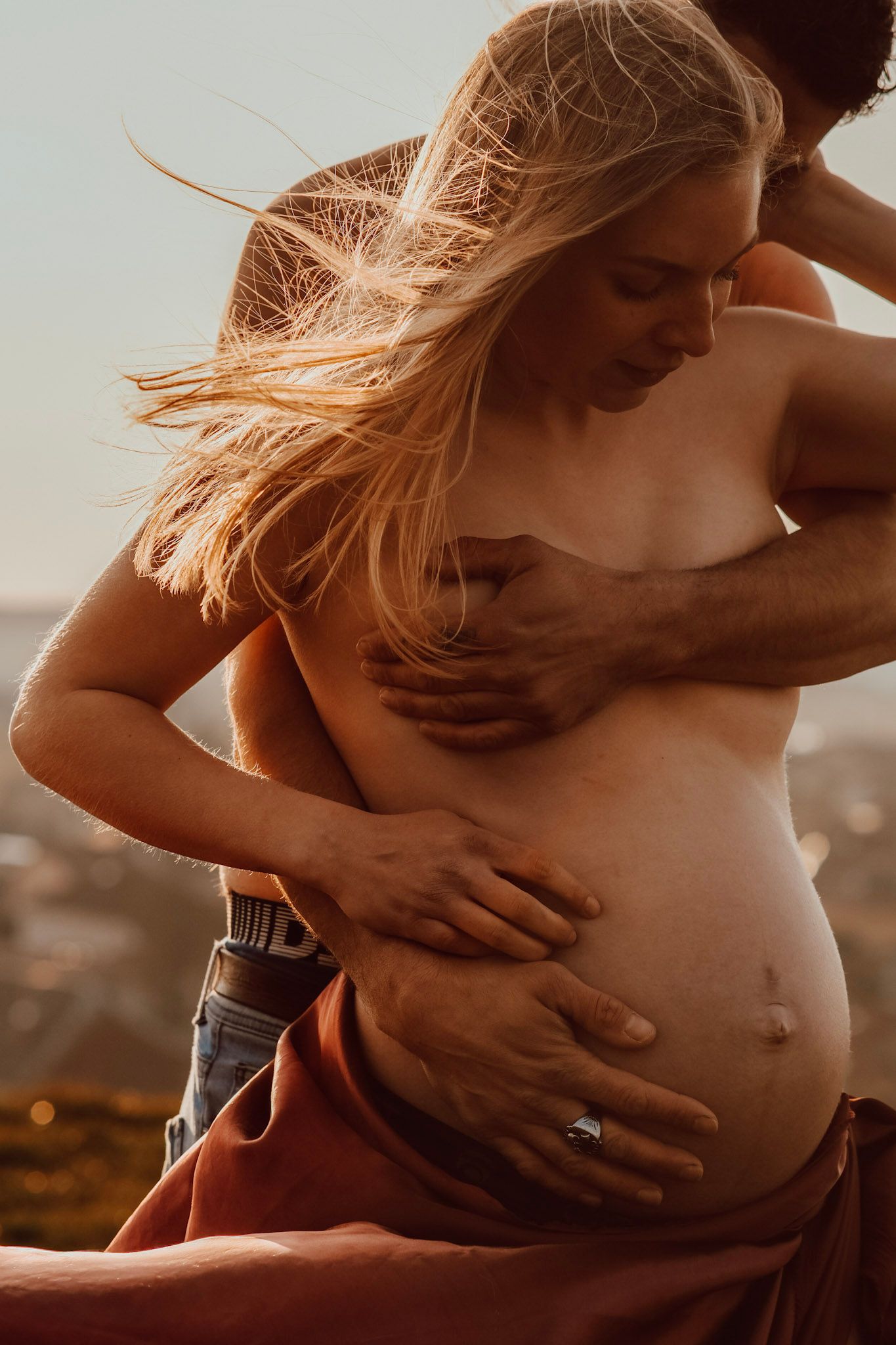 A pregnant woman and her partner share an intimate embrace outdoors at golden hour, with wind blowing through her blonde hair.