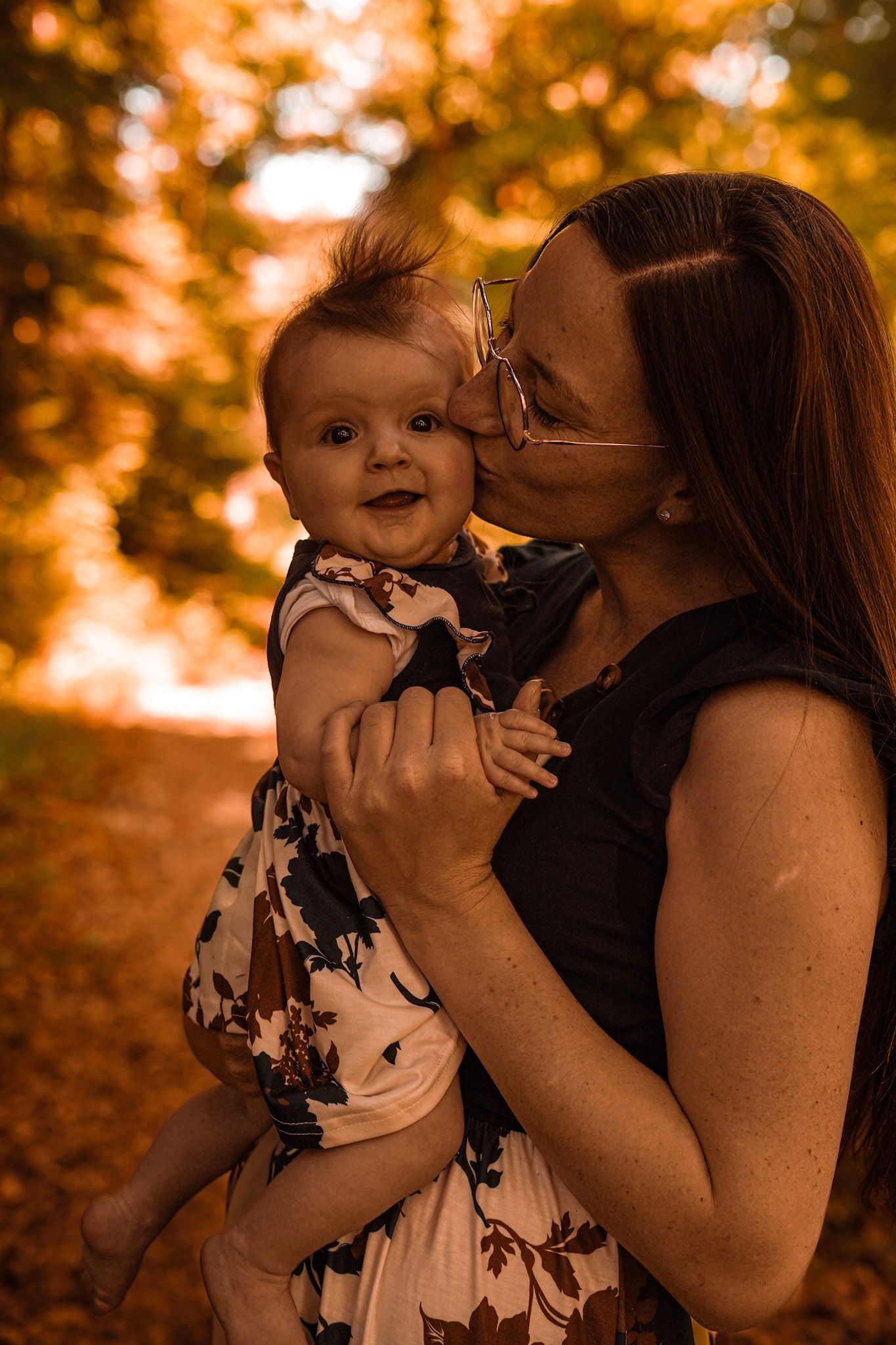 Mother kissing her smiling baby outdoors in autumn park with golden foliage background.