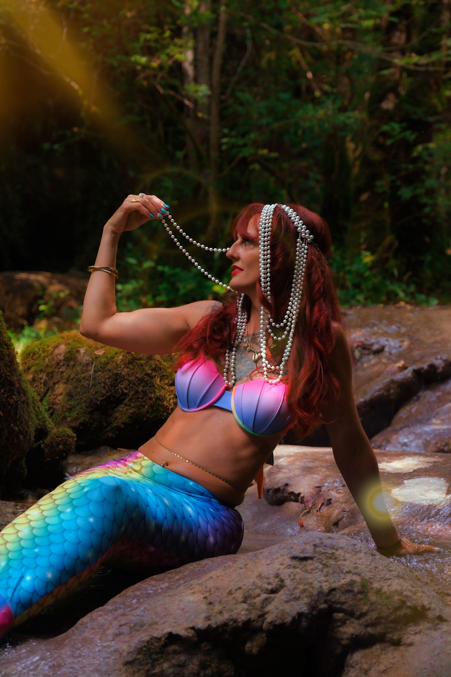 A woman in a colorful mermaid costume with rainbow tail and pearl jewelry poses on rocks in a forest.