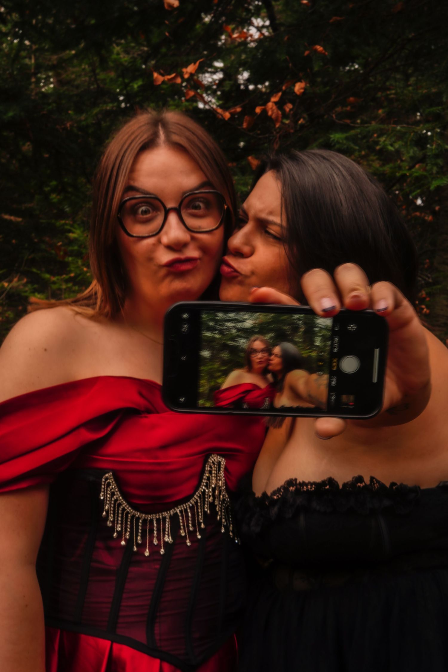 Two women in formal dresses taking fun selfie outdoors with trees in background.