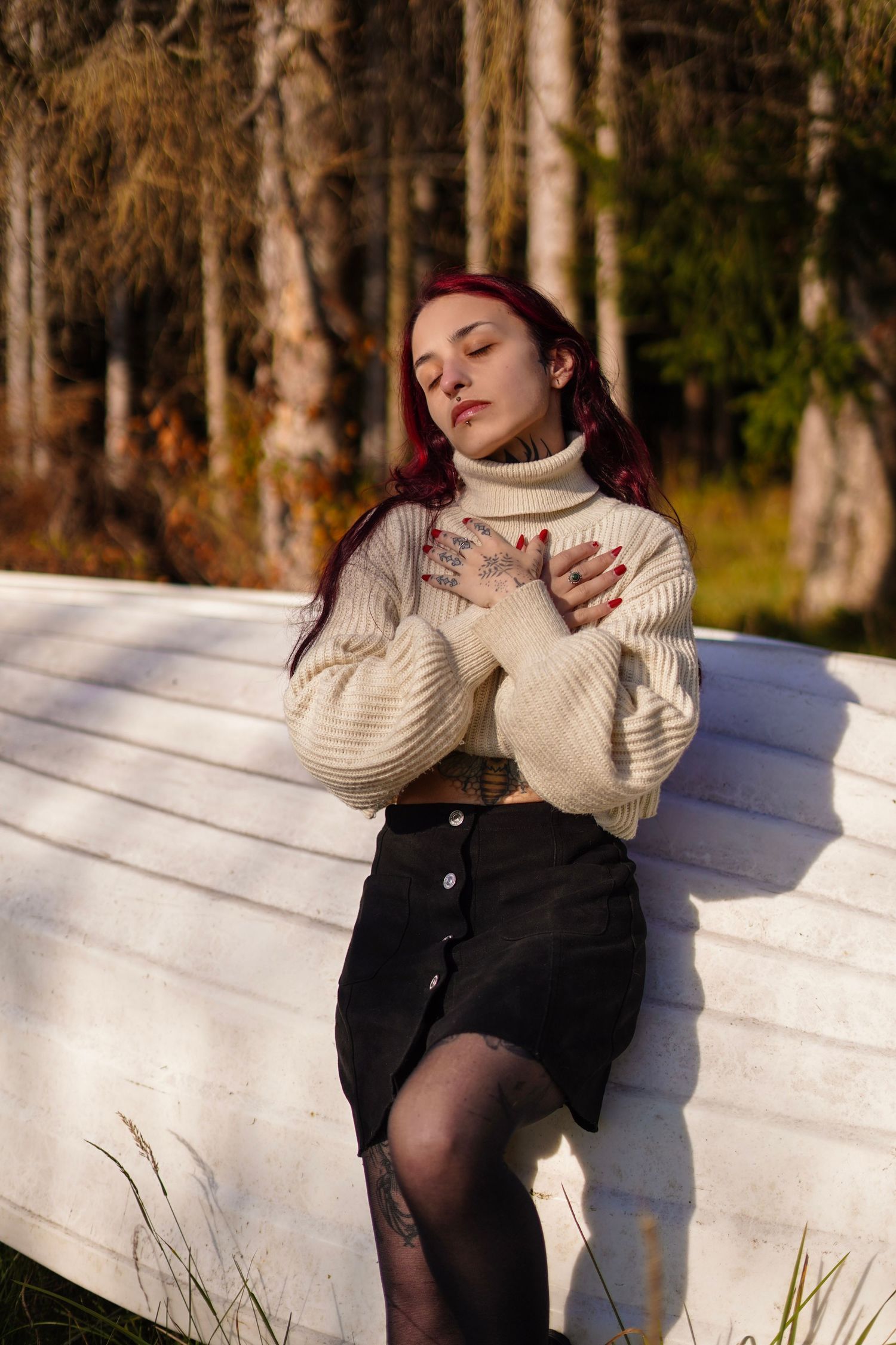 A red-haired woman in a cream turtleneck sweater and black skirt poses with eyes closed in sunlight.