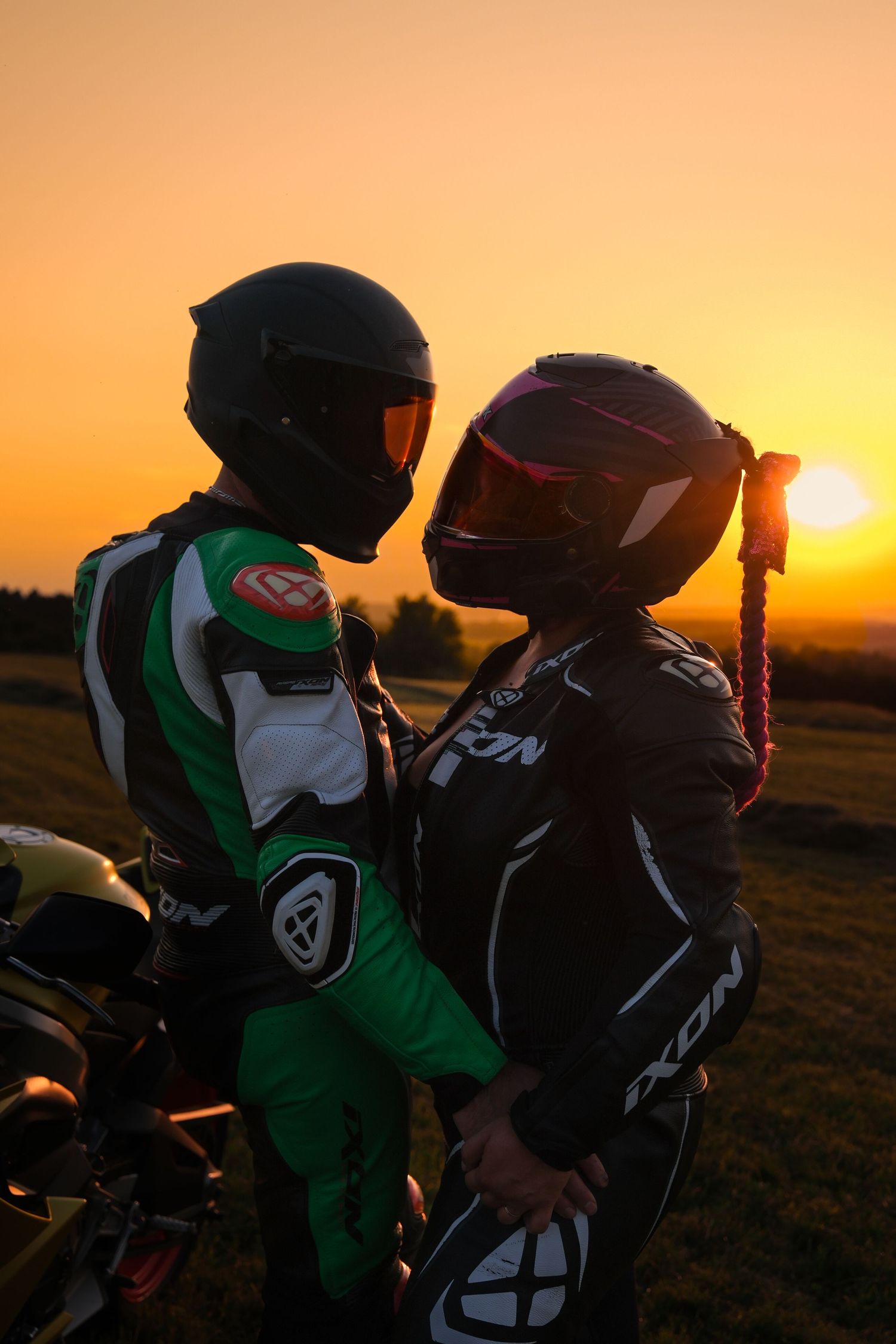 Couple in motorcycle racing suits sharing a romantic moment beside a bike at sunset.