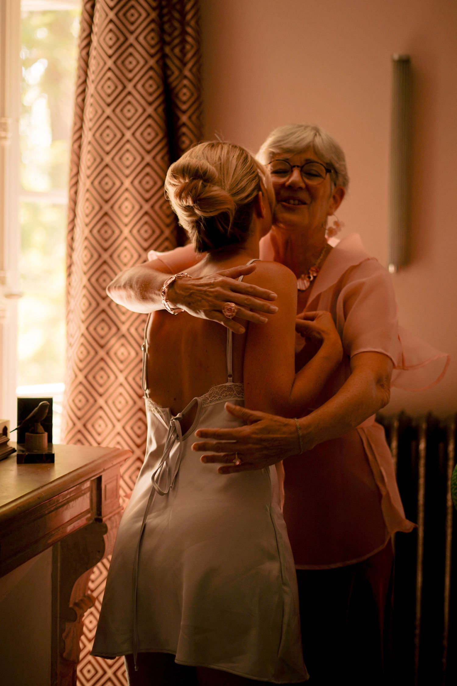 Tender moment as older woman helps bride in backless dress prepare for wedding day.