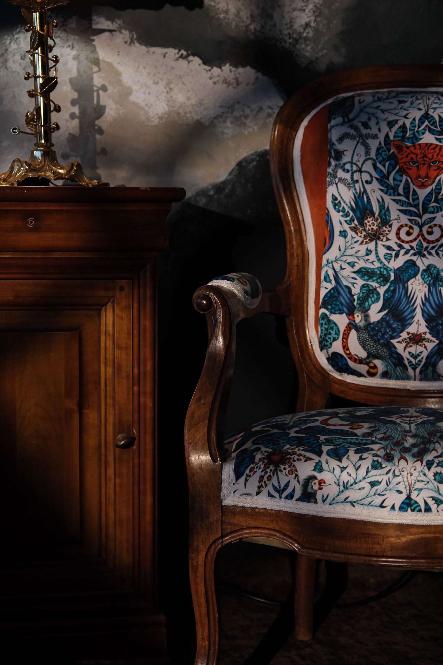 A vintage ornate armchair with floral upholstery beside a wooden cabinet.