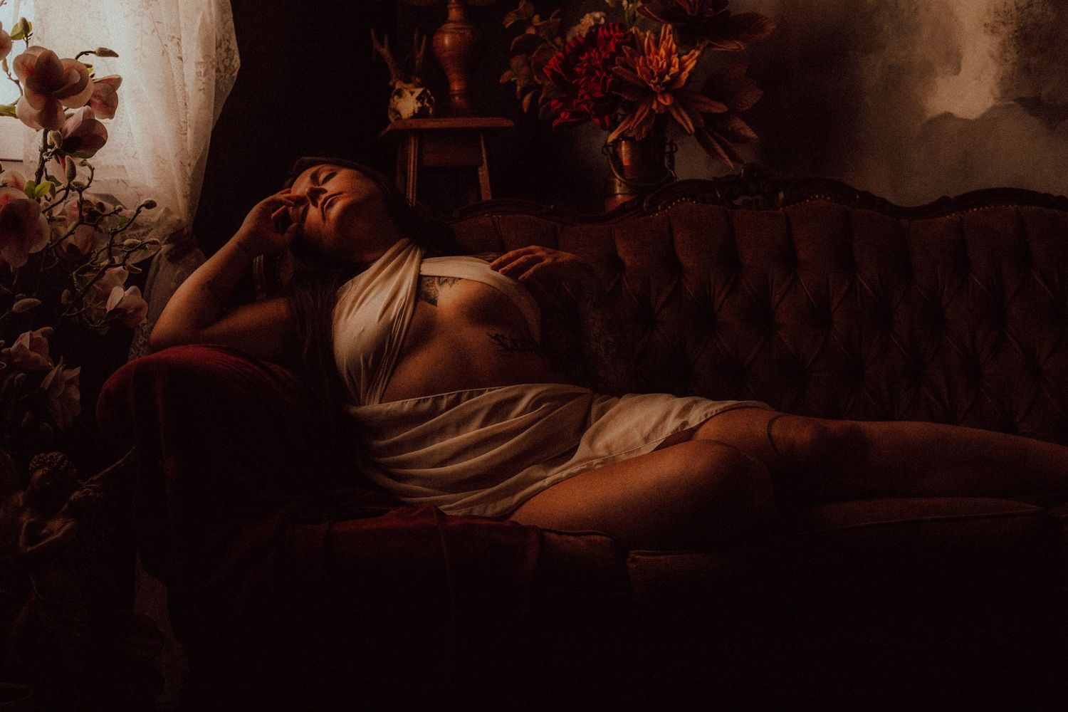 Woman in white lingerie reclining on antique velvet sofa surrounded by flowers in warm dim light.