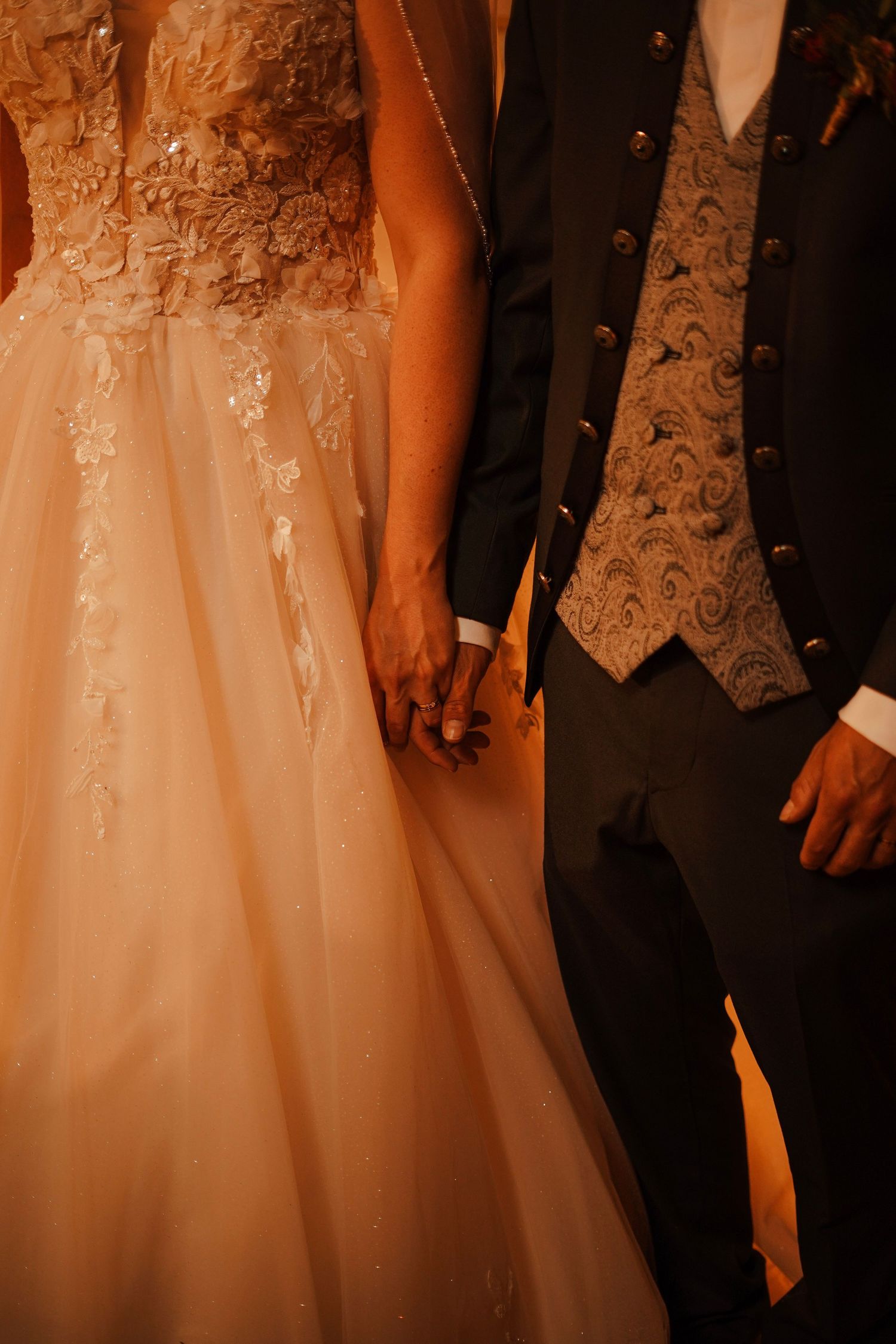 A bride and groom hold hands, showcasing an elegant lace wedding gown and ornate brocade vest in warm lighting.