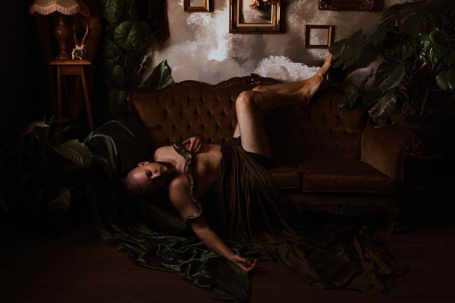 Woman draped in green silk fabric reclining on velvet sofa in dark moody botanical interior setting.