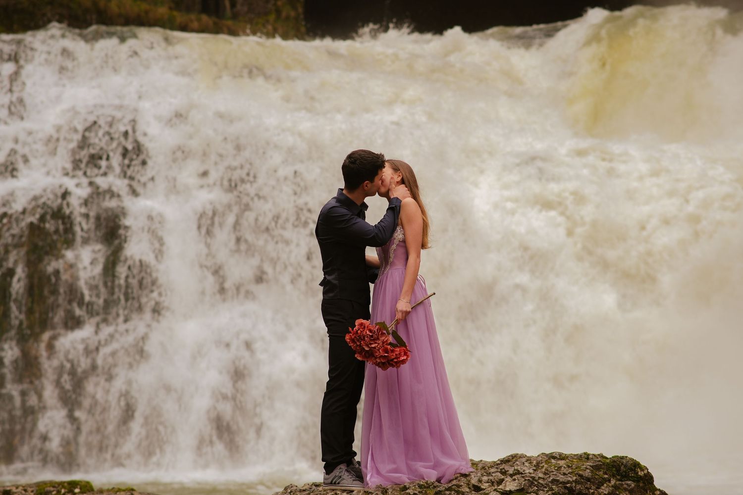Romantic couple kissing in front of a powerful rushing waterfall, woman in purple gown.