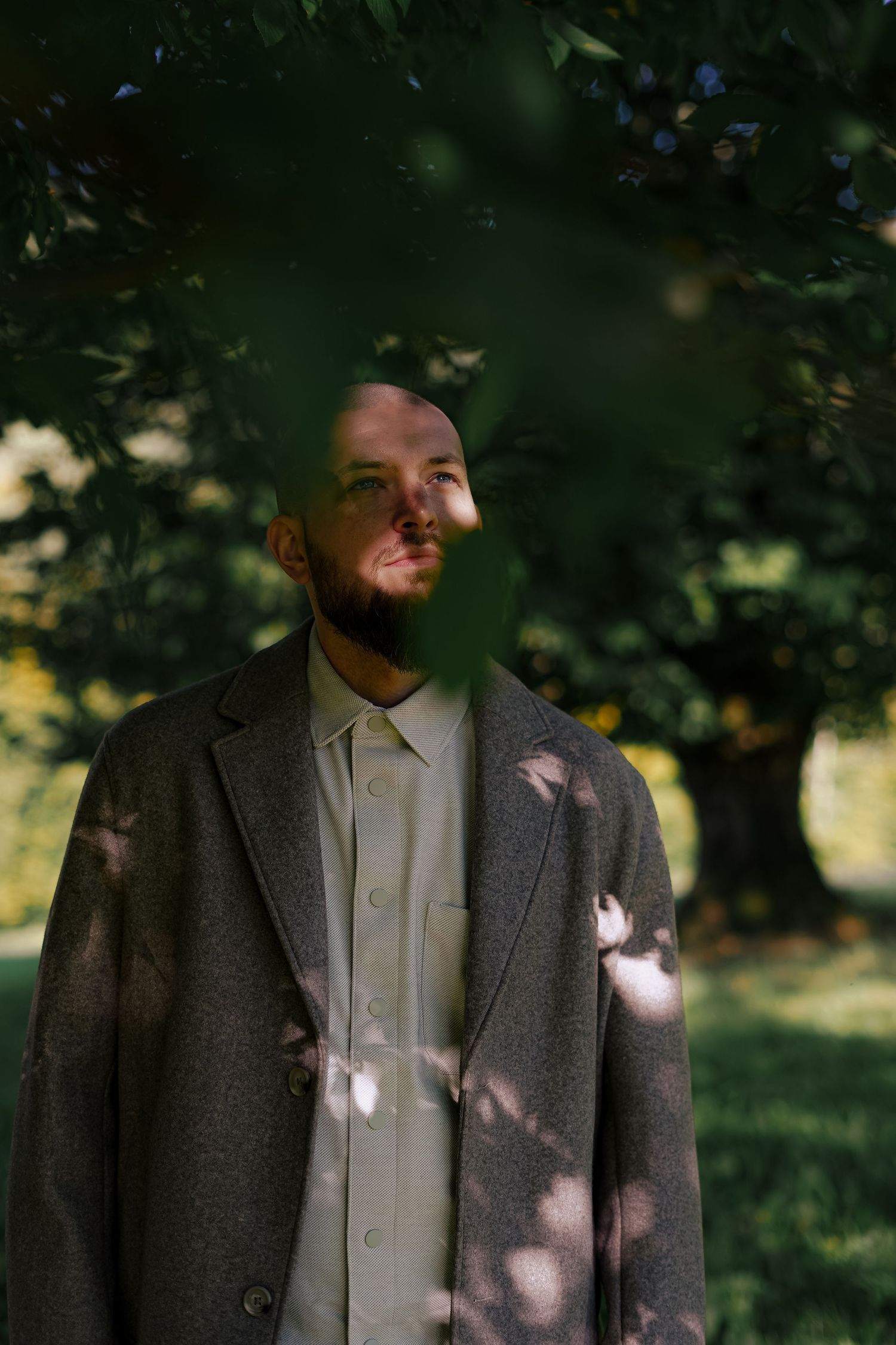Bearded bald man in gray blazer standing outdoors under green tree branches in sunlight.