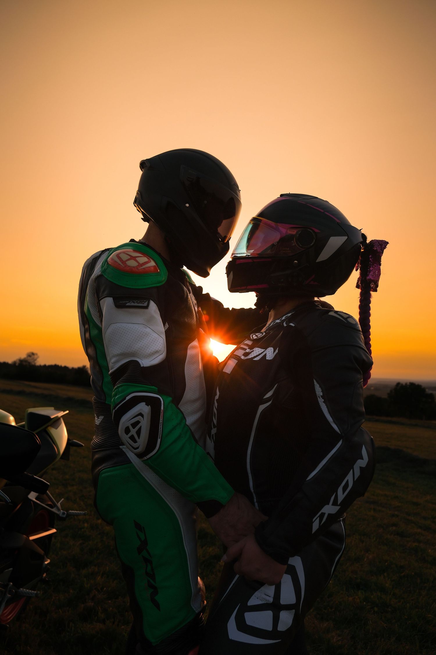 Couple in motorcycle helmets and racing suits sharing romantic moment at vibrant sunset.