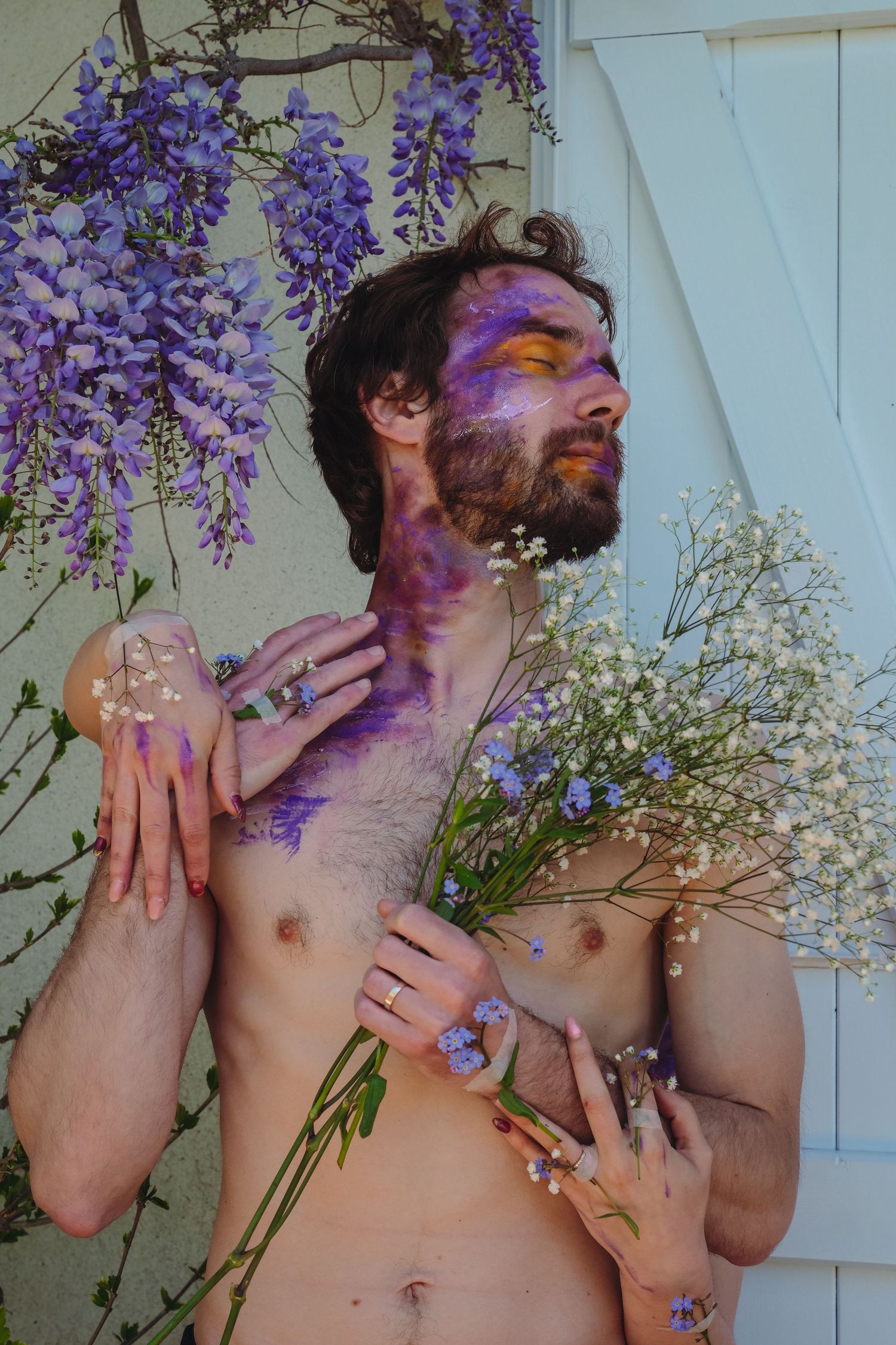 Shirtless bearded man with purple paint and flowers held by feminine hands outdoors.