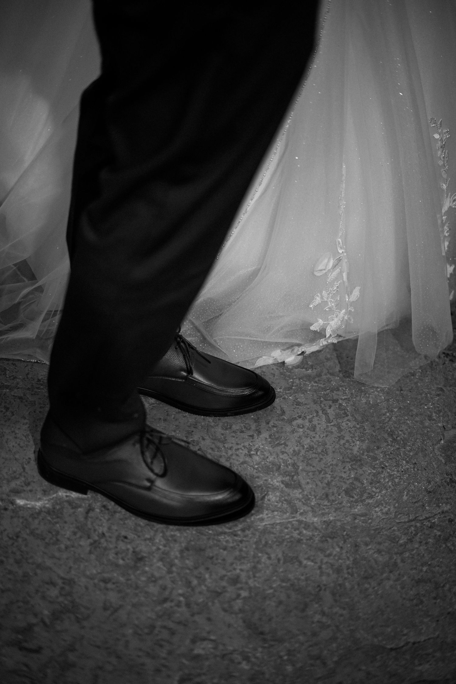 Black and white close-up of groom's leather dress shoes beside bride's white lace wedding gown.