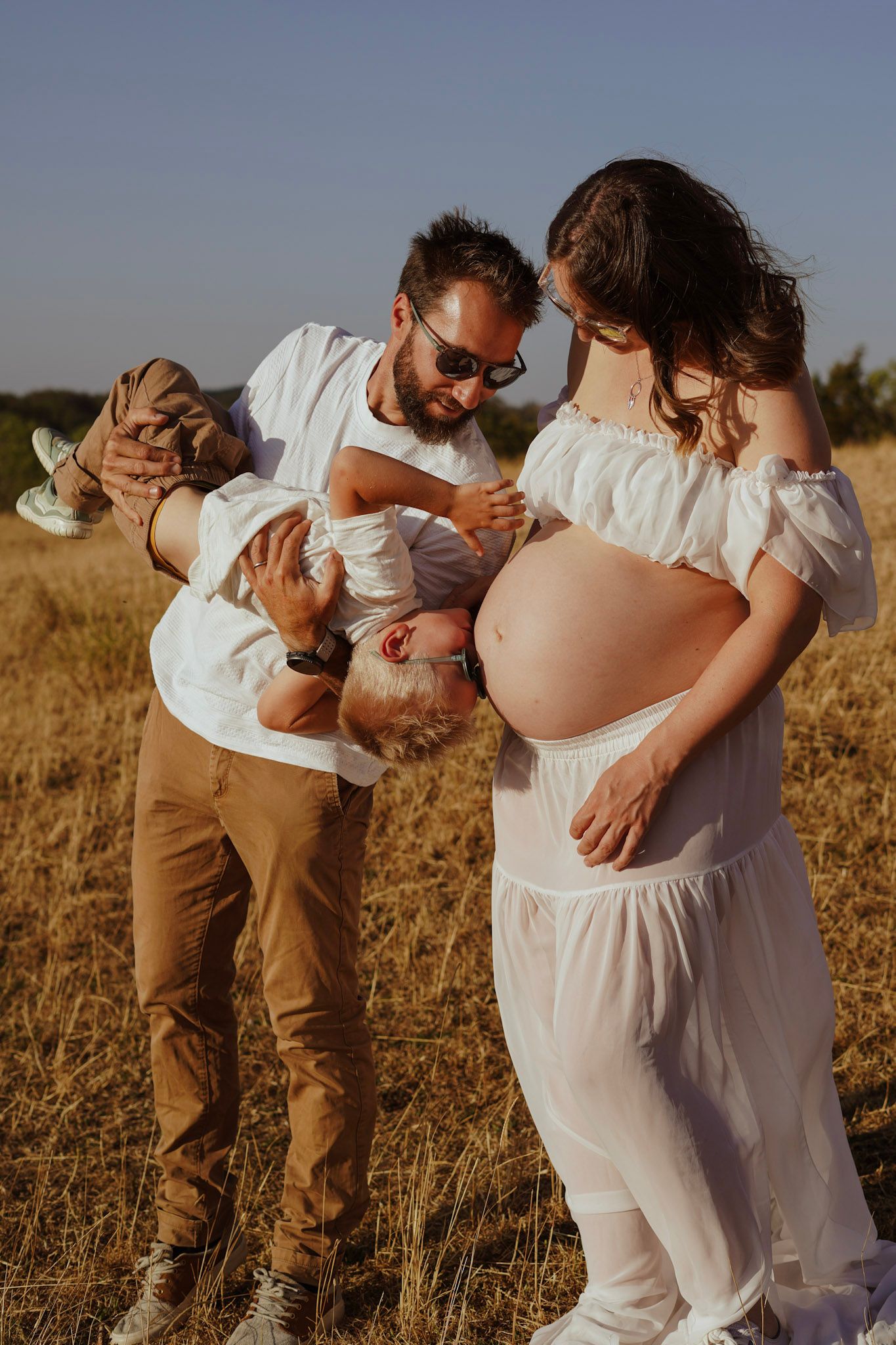 Pregnant woman in white dress with husband holding toddler upside down in golden field during sunny day.